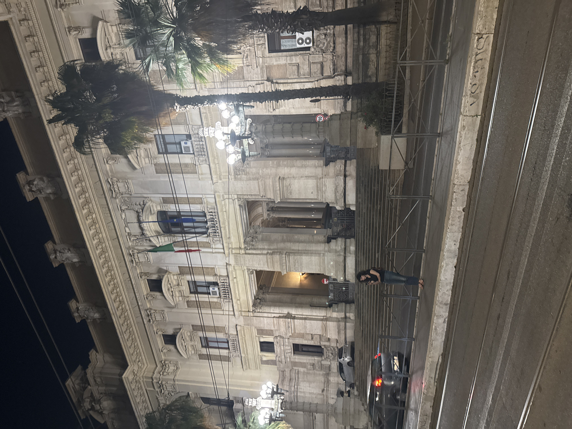 Here is a detailed description of the image:
The image shows a building facade in Rome, Italy, likely photographed at night. The architecture is ornate and classical, with multiple windows, decorative molding, and several palm trees. Two flags are hanging from the building; one is the flag of Italy, and the other appears to be the flag of the European Union.
On the right side of the image, there is a staircase structure made of metal railings and steps. A person is standing in front of the metal railing. On the street level, there are cars and streetlights. The road has tram tracks.