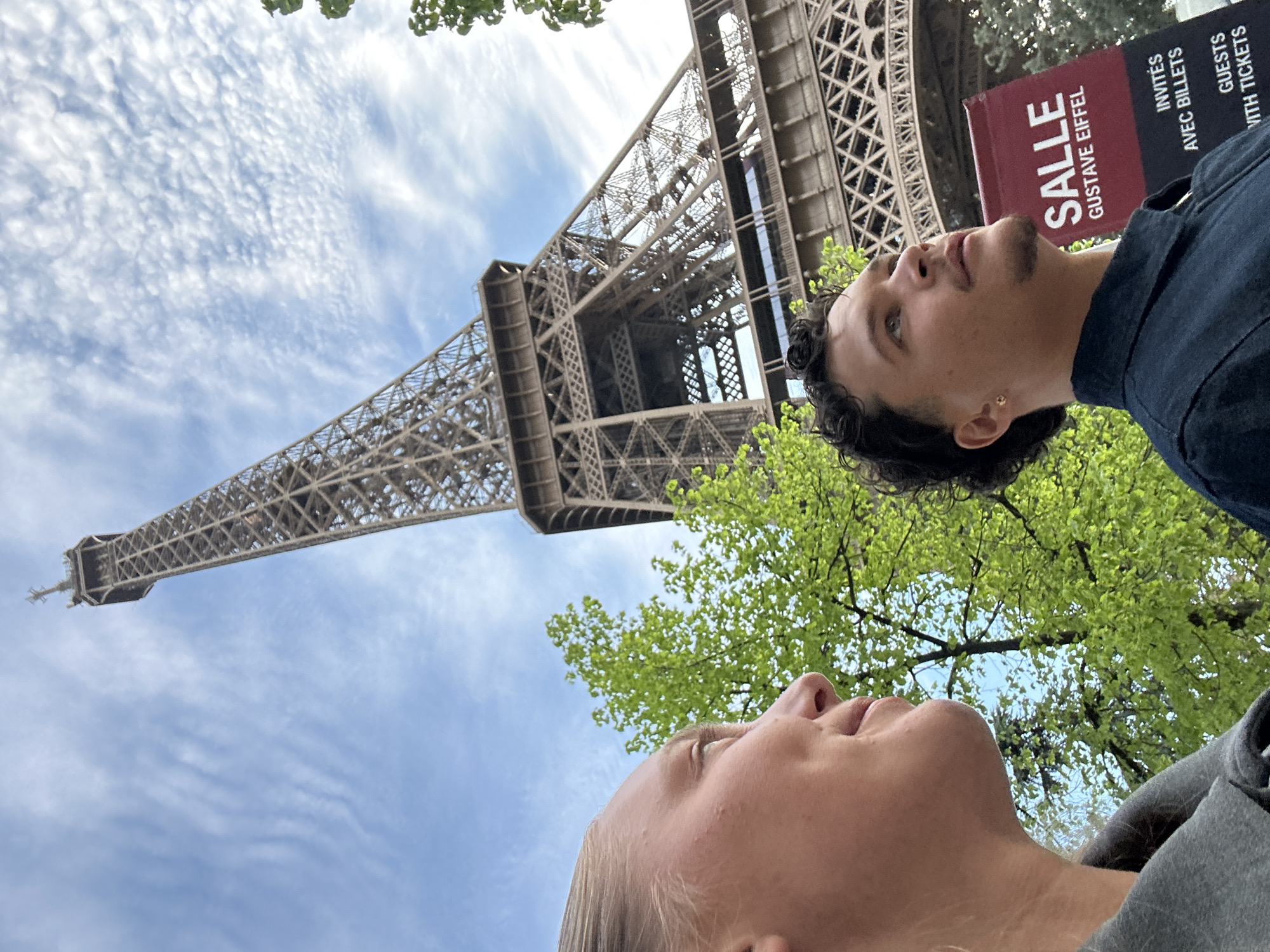 A low-angle shot in Paris, France, shows a woman and a man standing beneath the Eiffel Tower. The tower looms upward, its lattice structure prominent against a sky dotted with clouds. Lush green trees frame the edges of the tower. A sign reading "SALLE GUSTAVE EIFFEL" is visible near the man. The woman, in the foreground, is looking up, while the man is angled to the right.