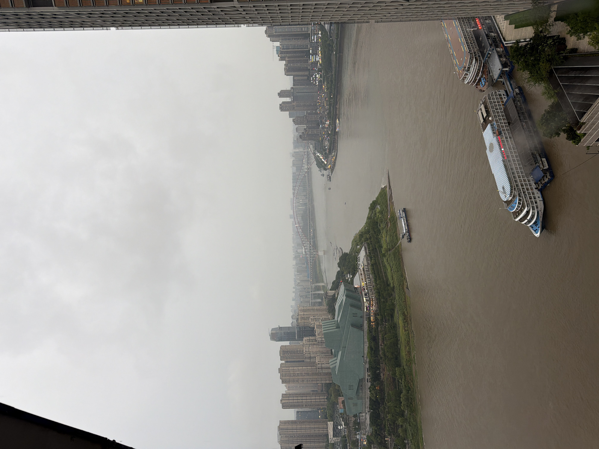A wide-angle, high-angle shot captures an overcast urban landscape along a wide, muddy river. On the left bank, a distinctive, multi-faceted green building sits at the edge of a cluster of tall, beige apartment buildings. Beyond these structures, a modern suspension bridge spans the river, connecting to a dense cityscape of skyscrapers on the opposite bank. Several boats, including two large passenger vessels docked together in the foreground and smaller boats further upriver, navigate the waterway. The scene is located in the Yuzhong District, China, a central area known for its vibrant riverfront and modern architecture. The overall mood is somber due to the overcast sky, suggesting a potentially humid or rainy day. No people are visible in the frame.