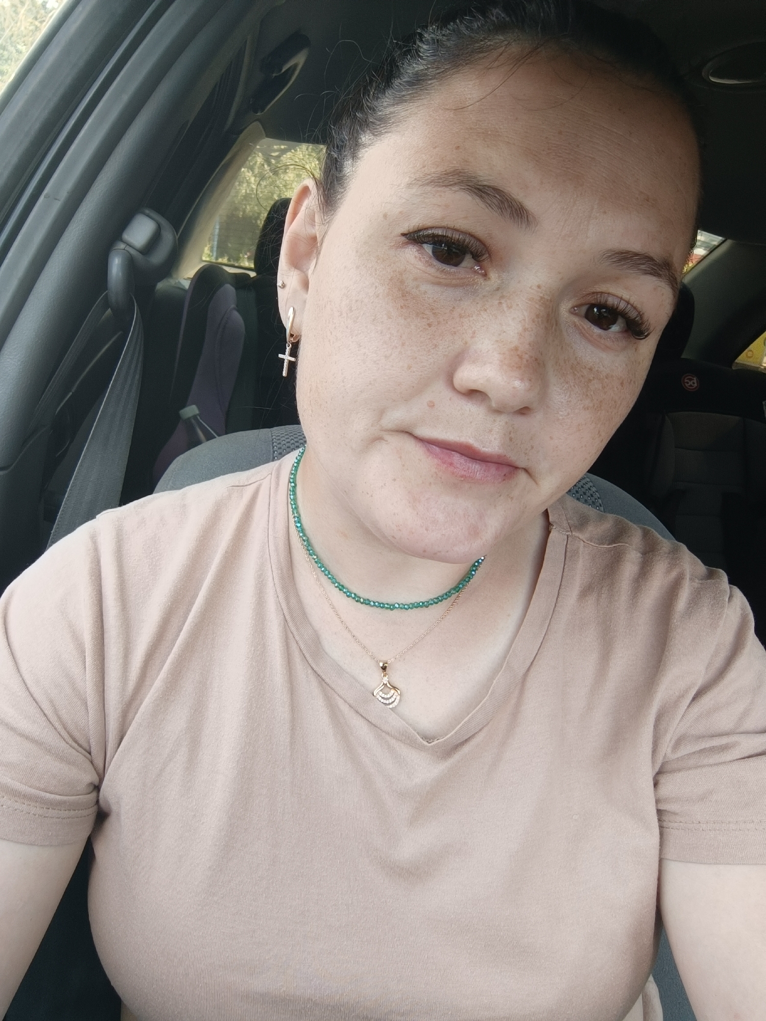 A medium shot captures a woman in a car, looking directly at the camera. She has dark hair pulled back and prominent freckles across her face. She is wearing a light brown t-shirt, a green beaded necklace, and a gold pendant. A cross earring is visible in her left ear. The background shows the interior of a car, with a seatbelt visible. The image does not provide enough information to determine a specific location in Chaikovsky, Russia.