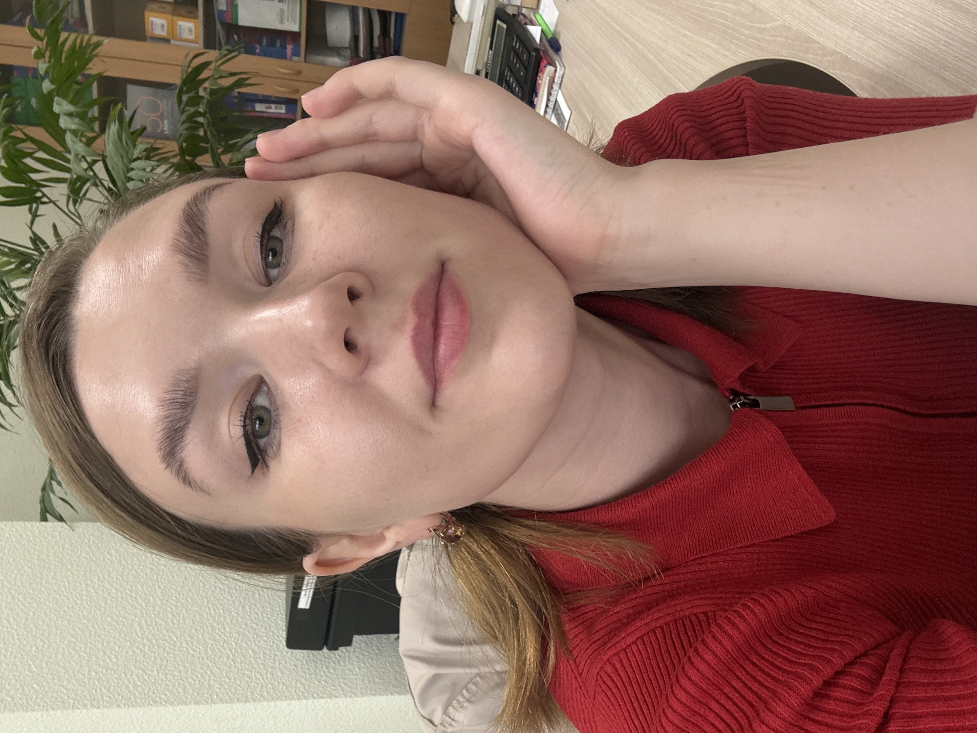 A woman with fair skin and dark eyeliner is facing the camera, her head tilted, resting her hand on her forehead. She is wearing a red, ribbed, collared shirt, zipped up to the collar. She has light brown hair, and her face is in focus. In the background, there is a plant with green leaves, a wooden cabinet with various items on its shelves, and a light-colored wall. The exact location is not discernible from the image. There is no evidence that the image was taken in Izhevsk, Russia.