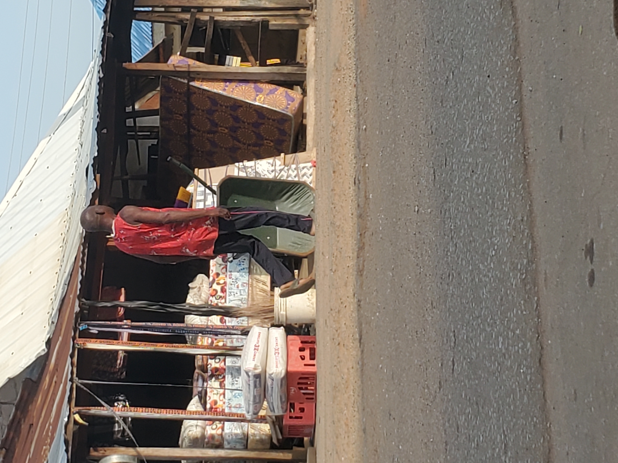 An outdoor shot shows a man in a red top and dark pants relaxing in a green wheelbarrow in front of a shop in Mararaba, Nigeria. The shop displays goods, including packages labeled "Biscuit 3X Great," stacked on wooden shelves and in red plastic crates. Above the shop, a mattress with a brown and gold patterned cover is stored. The location is on a paved street, with a concrete wall visible on the right side of the frame. The sky is light, suggesting it is daytime.