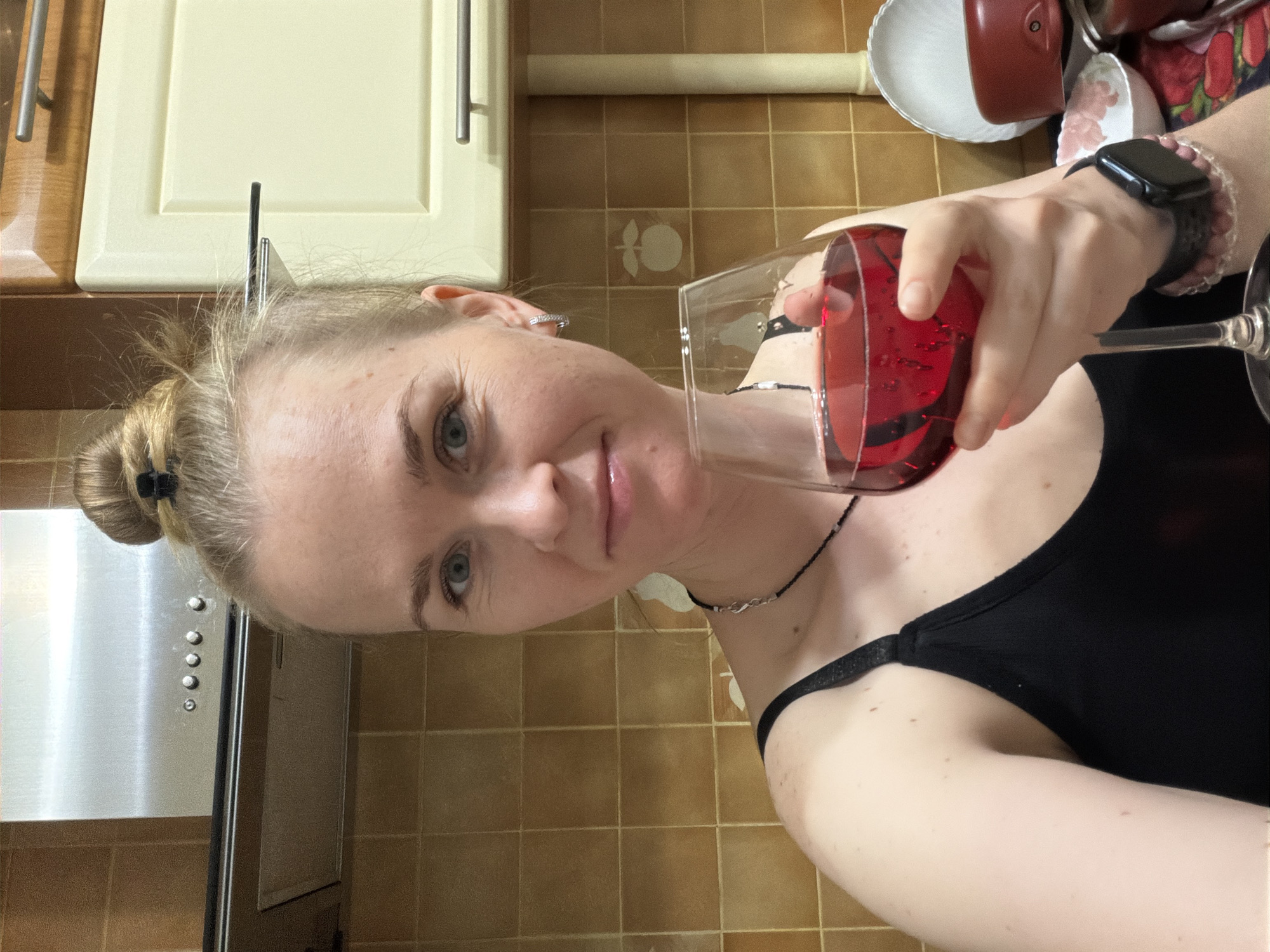 A young Caucasian woman with blonde hair styled in a bun is holding a glass of red wine and taking a selfie in what appears to be a kitchen. She is wearing a black tank top and a necklace. The background shows a tiled wall, a kitchen range hood, and cabinets. The overall lighting suggests an indoor setting, possibly during the day or evening. The location is not determinable from the image itself.