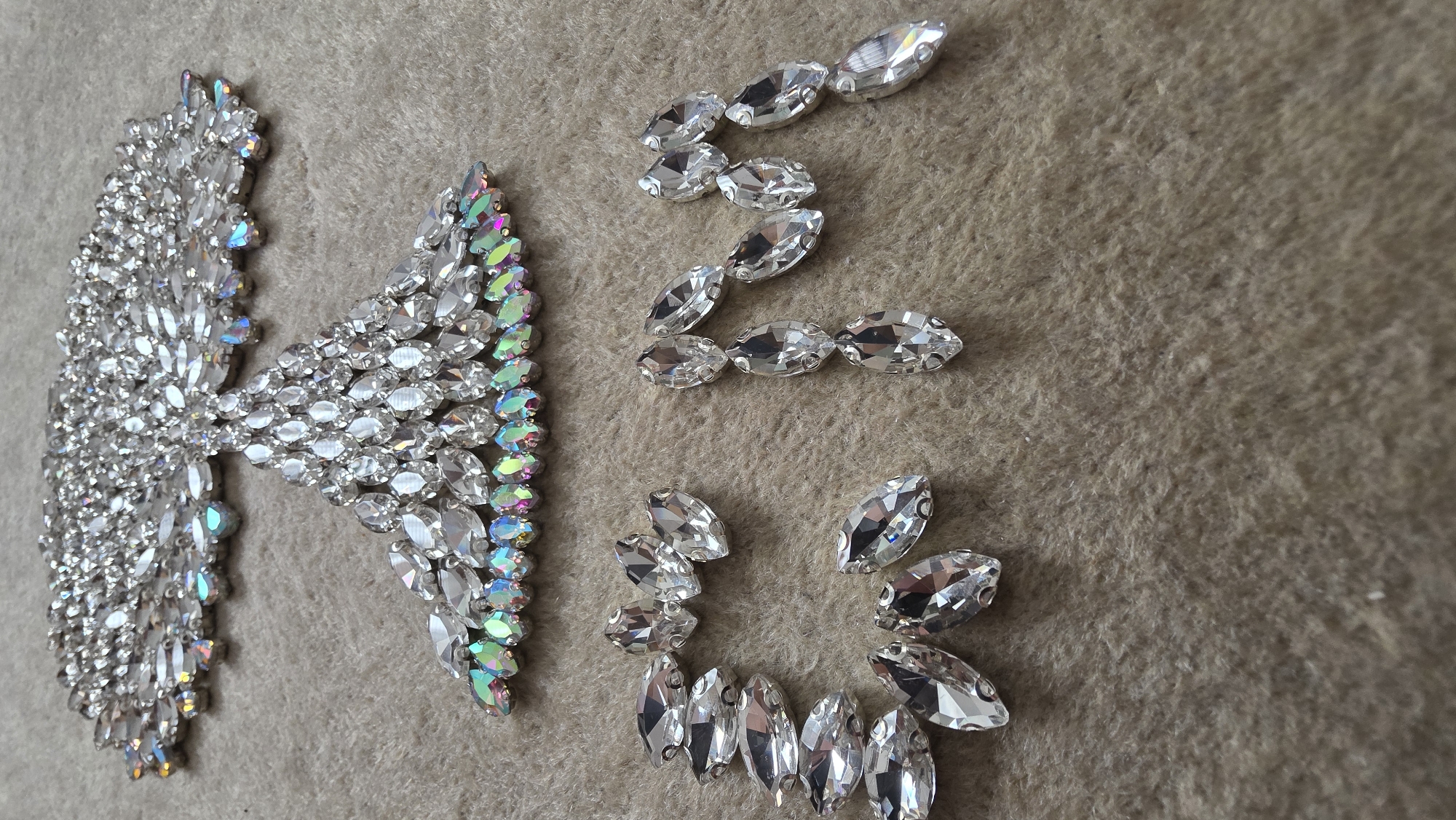 The image shows a flat lay arrangement of various clear, faceted rhinestones on a beige, textured surface. The rhinestones are organized into distinct shapes: a large, dense, butterfly-like design, an angular arrangement resembling the letter "Z," and a partial circular arc. No people are visible. The location is not discernible from the image. The only objects are rhinestones of varying shapes and arrangements, and the beige surface upon which they rest. No actions or interactions are depicted.