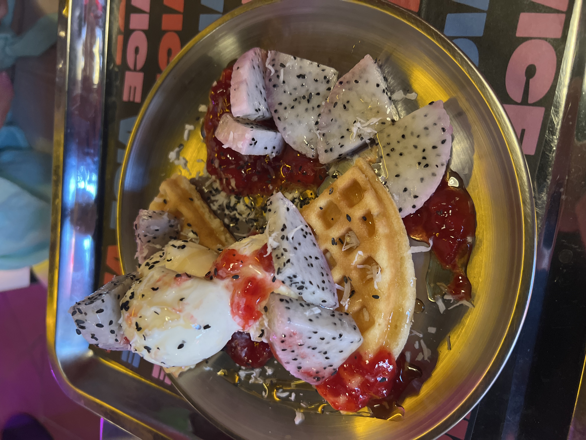 A close-up, high-angle shot captures a vibrant dessert served on a golden-hued metallic plate, likely in an indoor setting like a cafe in Bac Nha Trang Ward, Vietnam. The dish features several components artfully arranged.
The main subjects are pieces of golden-brown waffles, some showing their characteristic grid pattern, accompanied by numerous triangular wedges of white-fleshed dragon fruit, speckled with tiny black seeds. Nestled among these are scoops of a creamy white substance, likely ice cream or frozen yogurt, which appears slightly melted.
The dessert is generously adorned with a bright red, chunky sauce, possibly a berry jam or compote, and a glistening clear syrup or honey drizzled over the entire ensemble. A scattering of black sesame seeds and some white shredded flakes, possibly coconut, adds further texture and visual interest.
The metallic plate has a reflective surface, mirroring ambient light. It rests on a darker surface, possibly a tray or table, which also shows some reflections. In the background, partially visible, is a dark sign or wall graphic with distinct lettering. The text "ICE" is visible vertically in pink, and parts of the word "LOVE" (with the "L" partially obscured) are visible horizontally in pink and blue lettering. The overall lighting suggests an indoor, possibly evening, ambiance. There are no people visible, and no specific activity is taking place other than the presentation of the dessert.