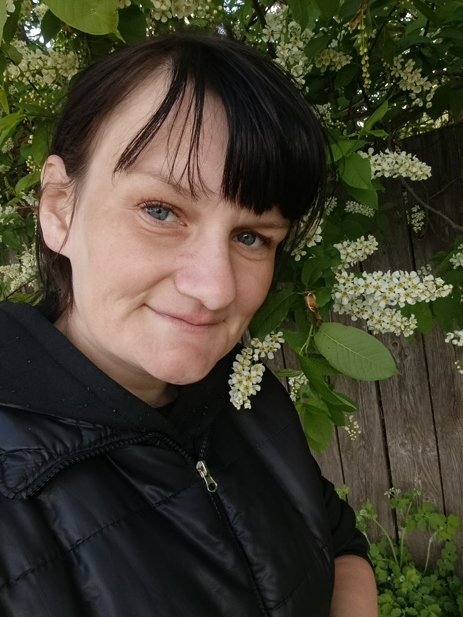 The image shows a woman with short, dark hair and blue eyes, smiling at the camera. She is wearing a black jacket. Behind her is a wooden fence with white flowers blooming on a bush or tree. The location is Фоки, Russia.