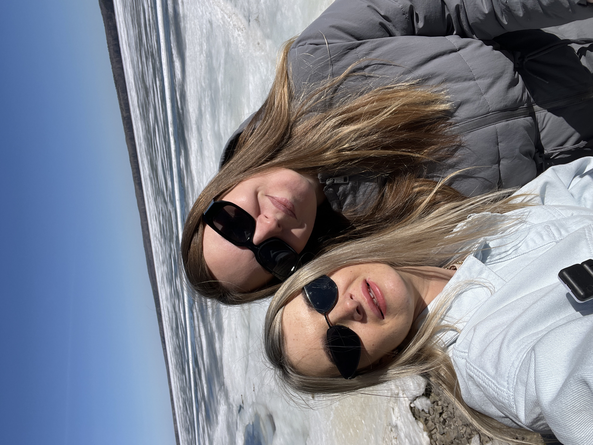 Two women lie close together on a frozen surface, potentially a body of water. The woman on the left has long brown hair and wears a gray puffer jacket and black sunglasses. The woman on the right has long blonde hair and wears a light blue denim jacket and black sunglasses. Behind them, the image reveals a stark contrast between a frozen, possibly snowy or icy, surface and a clear blue sky. Given the request to provide information pertinent to Chaikovsky, Russia, it is possible they are positioned on or near the Kama River or one of the other water bodies surrounding the city, frozen in the winter season.