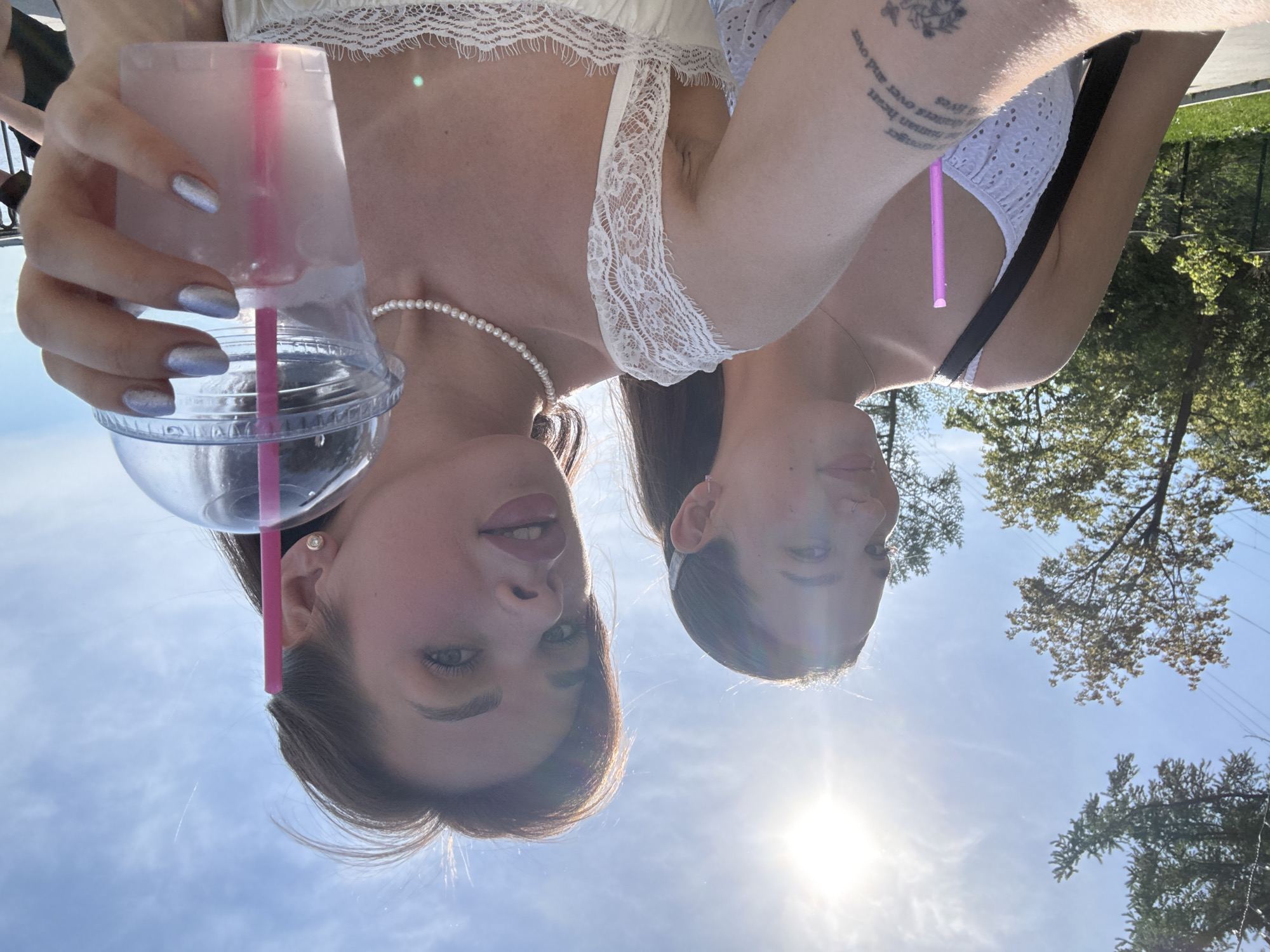 Two women are posing for a selfie outdoors. The woman on the left is holding a clear plastic cup with a pink straw. She is wearing a white top and a pearl necklace. The woman on the right is wearing a white top with a black strap. There is a tattoo on her upper arm. The sky is visible in the background, along with trees and foliage. The picture was taken in Perm, Russia.