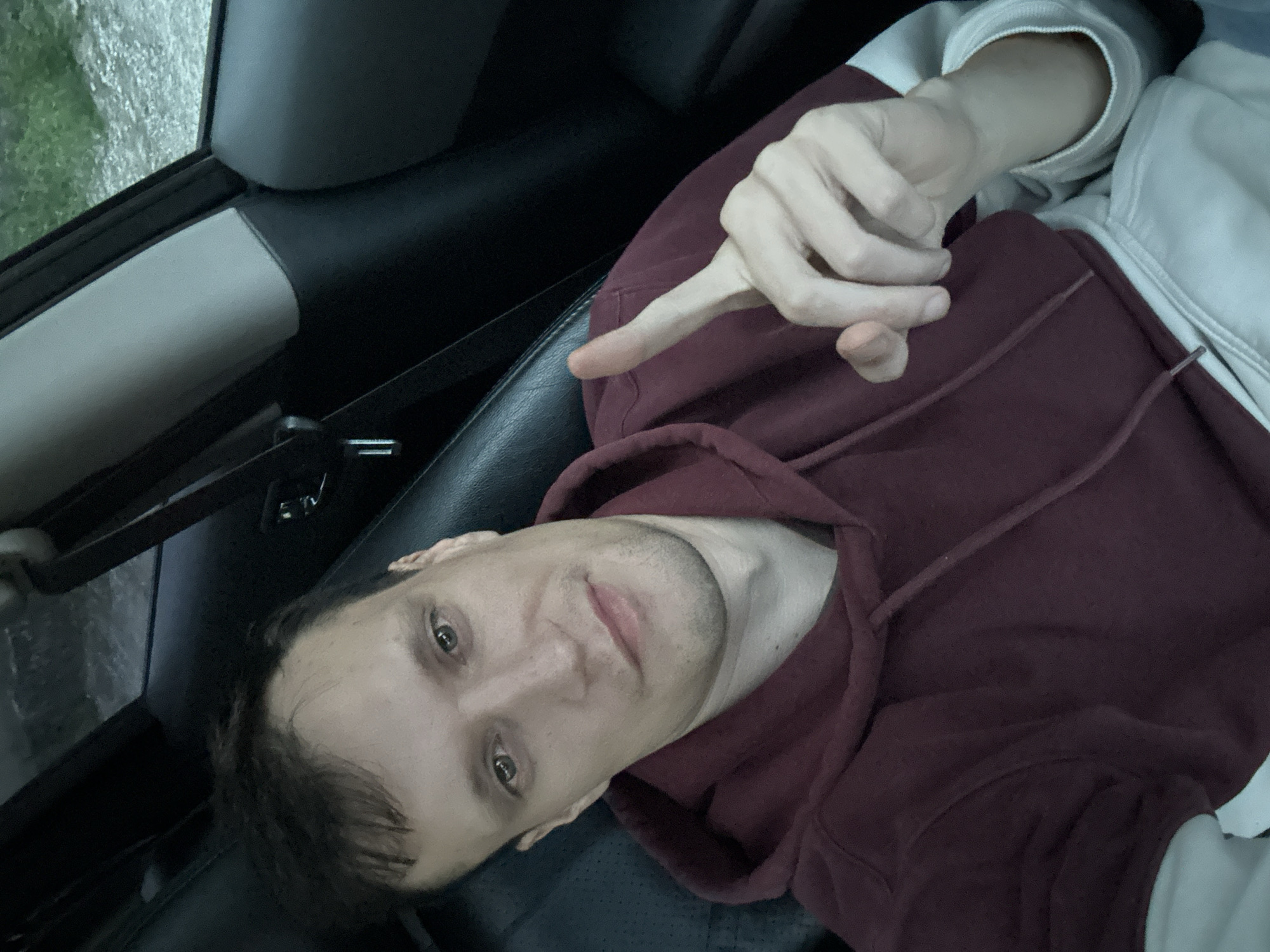 A man with light skin and short brown hair is in the passenger seat of a car. He is wearing a maroon hoodie over a white long-sleeved shirt. He is looking at the camera and holding his right index finger up. The car's interior, including black leather seats and a gray pillar, is visible. The scene appears to be in a rural area with trees and gravel visible through the car window. Shatava, Ukraine is not depicted in this image.