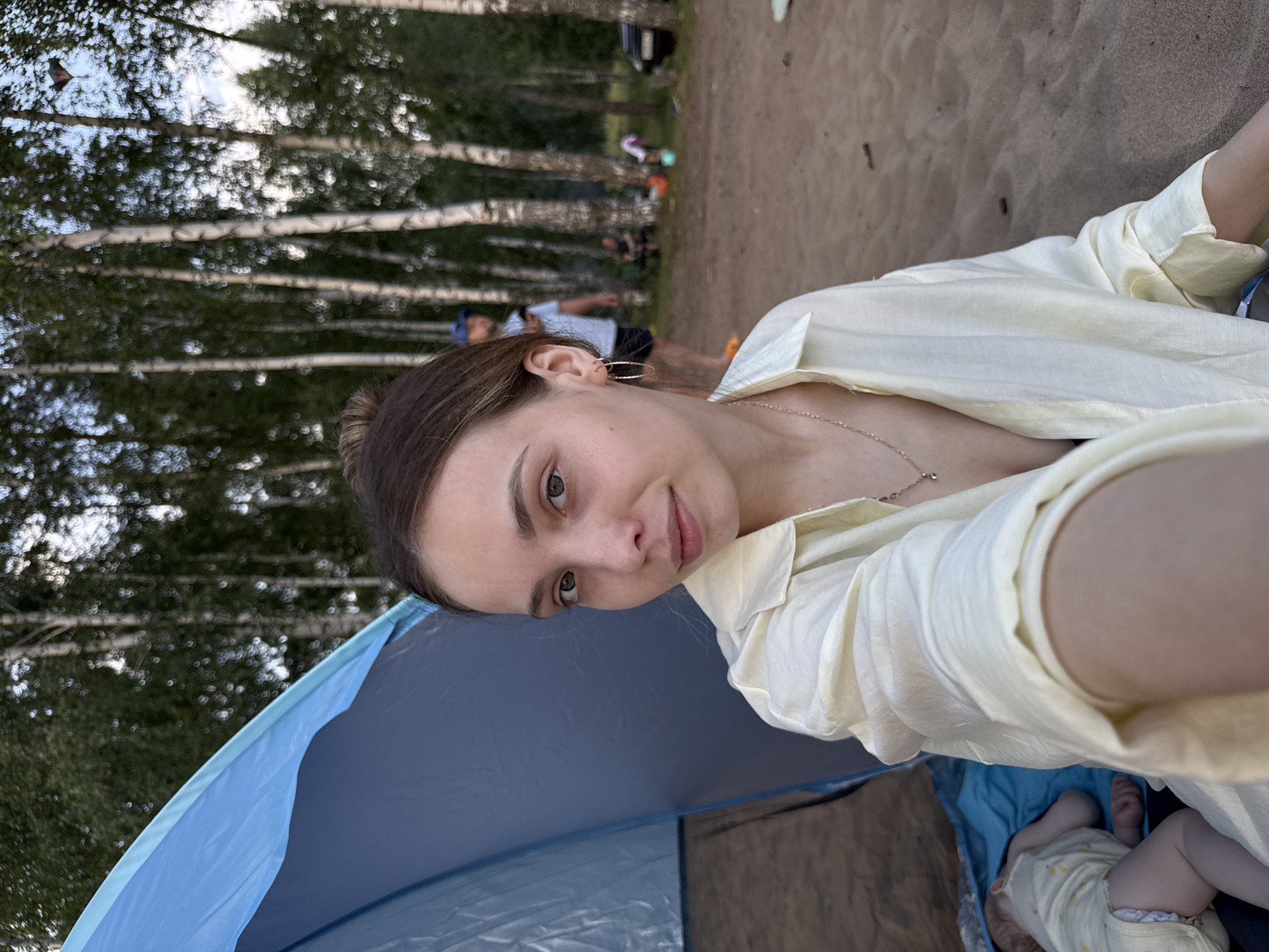 A medium shot captures a young woman in the foreground, seated on sandy ground beside a blue and grey tent. She is looking towards the camera with a neutral expression, her light-colored, collared shirt slightly open, revealing a necklace. Her hair is pulled up into a bun. To her left, a baby is visible, lying on a blue fabric within the tent. In the background, a grove of birch trees stands tall, with a few people milling about in the distance. One person, wearing a blue cap, is looking at their phone. Another person is seated near a campfire. A car is parked on the right side of the frame. The overall setting appears to be a park or a beach area, likely in Chaikovsky, Russia, given the presence of birch trees and the general atmosphere of a recreational outdoor gathering.