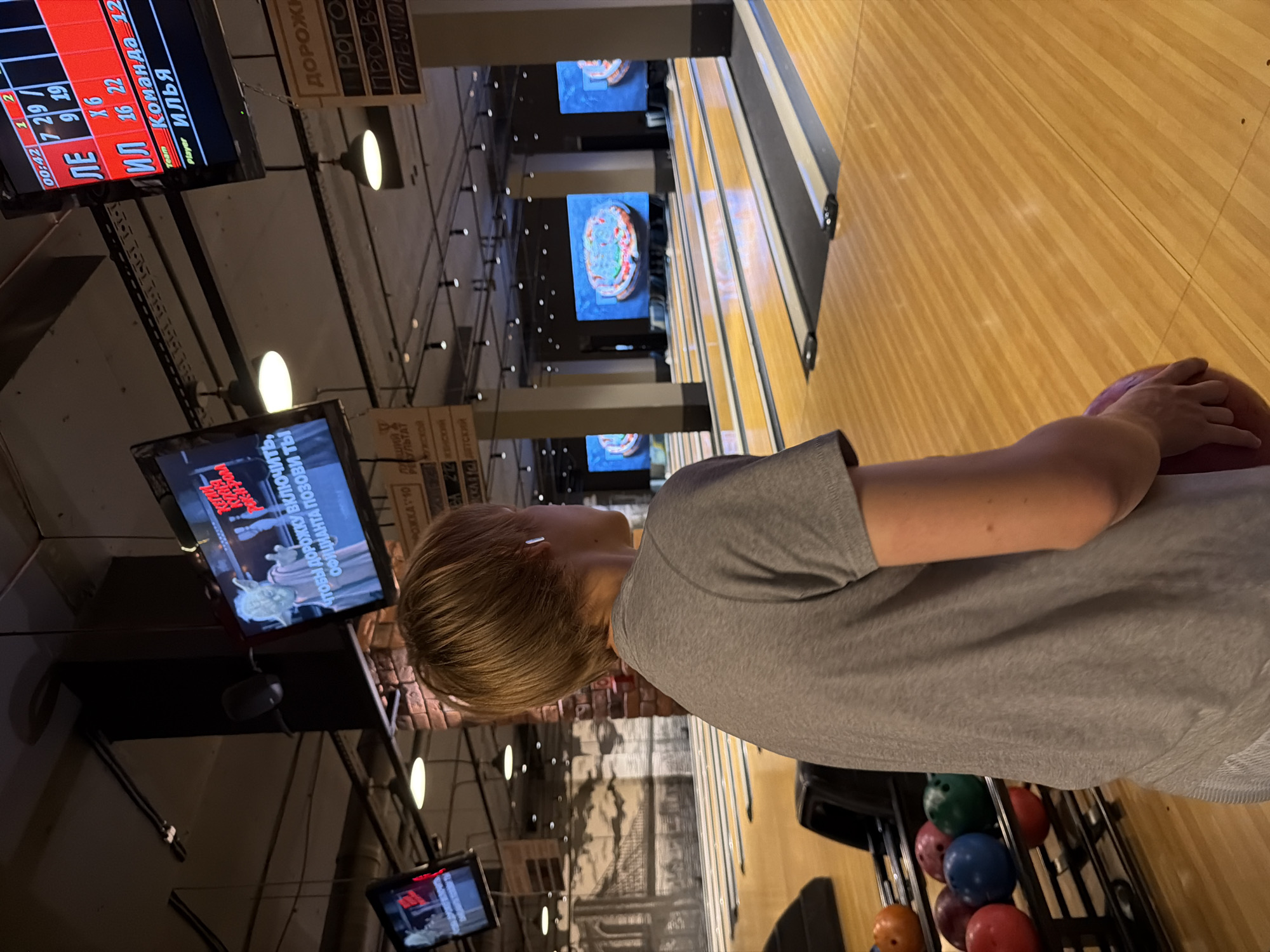 In a dimly lit bowling alley, a person is about to bowl. They are wearing a grey t-shirt and have short, blonde hair, with visible earbuds. They are holding a red bowling ball in their right hand, poised to throw it down the lane. The bowling alley features wooden lanes and overhead screens displaying scores and animations. The atmosphere appears to be recreational, possibly a casual game or competition.
Visible text on screens and signage includes Russian words related to bowling, such as "Команда" (Team), "Player", and lane numbers. One screen displays a timer at "00:42". Another screen shows "ЧТОБЫ ДОРОЖКУ ВКлючить, ОФИЦИАНТА ПОЗОВИ Ты" which translates to "To turn on the lane, call the waiter". Signs with Russian text like "РОЖКА 10", "РЕКУЛЬТАТ", "Мужской", "ДОРОЖН", "Просве", and "ГОРБУНО" are also visible, suggesting a Russian-speaking environment. The overall lighting suggests it is likely evening or indoors, with artificial lighting illuminating the scene. The setting is clearly a bowling alley, indicated by the lanes, balls, and scoring displays.