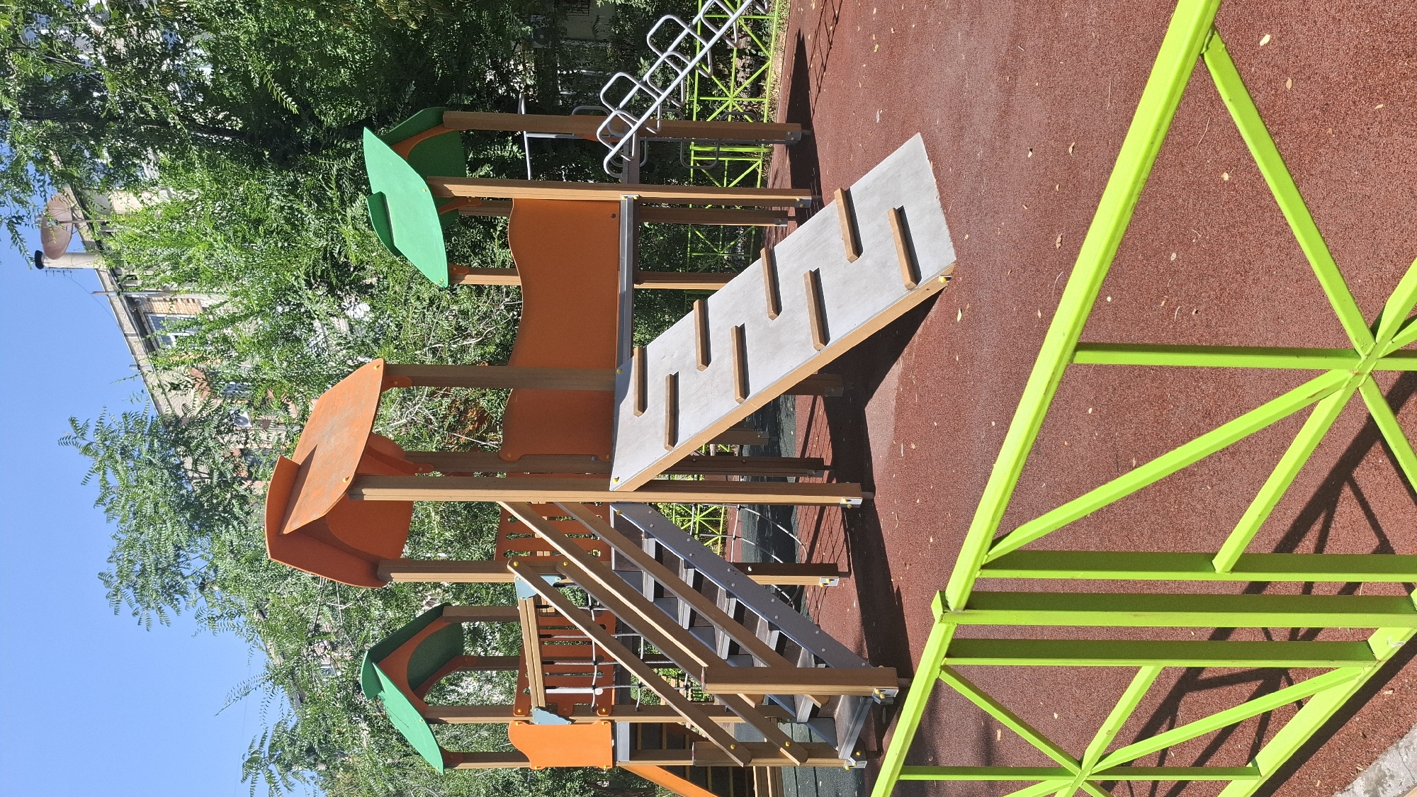 An outdoor playground is shown on a sunny day. The playground has a wooden and plastic structure with orange and green roofs. A ramp with wooden slats leads up to the structure, and a set of stairs is also visible. A bright green metal fence is in the foreground. The ground of the playground is covered in a red, rubberized material. In the background, there are trees and a multi-story building. The location is identified as Shymkent, Kazakhstan. There are no people present in the image.
