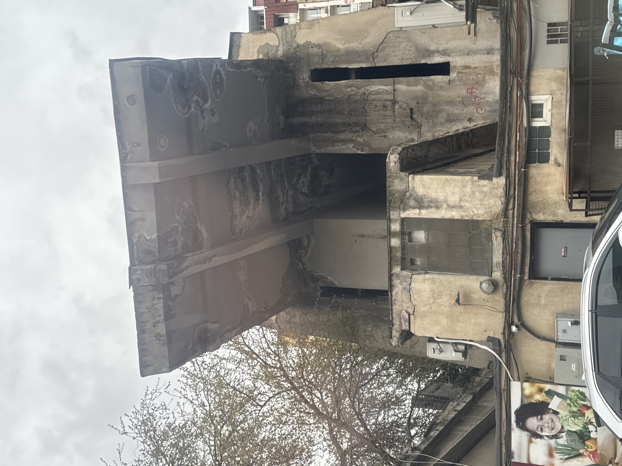The image displays a weathered building facade in an urban setting, likely Tbilisi, Georgia, under an overcast sky during daylight hours. The building exhibits significant signs of age and wear, with extensive peeling plaster, discoloration, and dark moisture stains visible across its concrete or stucco surfaces. Prominent concrete beams protrude from the main structure, also showing similar signs of deterioration.
Numerous black electrical cables and utility wires are haphazardly strung across the building's exterior. Several small, dark, rectangular openings are visible on the upper walls, along with a small, square, white-framed window featuring grilles and a section of glass block windows below it. A white air conditioning unit is mounted high on the facade.
In the lower right portion of the image, the front and side of a silver car are visible, parked close to the building. Adjacent to the car, a large advertisement poster features a smiling young woman holding an abundance of fresh produce, including broccoli, peppers, and green leafy vegetables. Metal utility boxes are affixed to the wall near the car, and a dark grey metallic door or gate is partially visible on the far right.
A tree with sparse, light-colored foliage is visible in the lower left foreground and through openings in the building structure, suggesting either early spring or late autumn. Faint red graffiti, possibly "JOXO," is visible on one of the walls. The scene captures an older, lived-in urban environment.