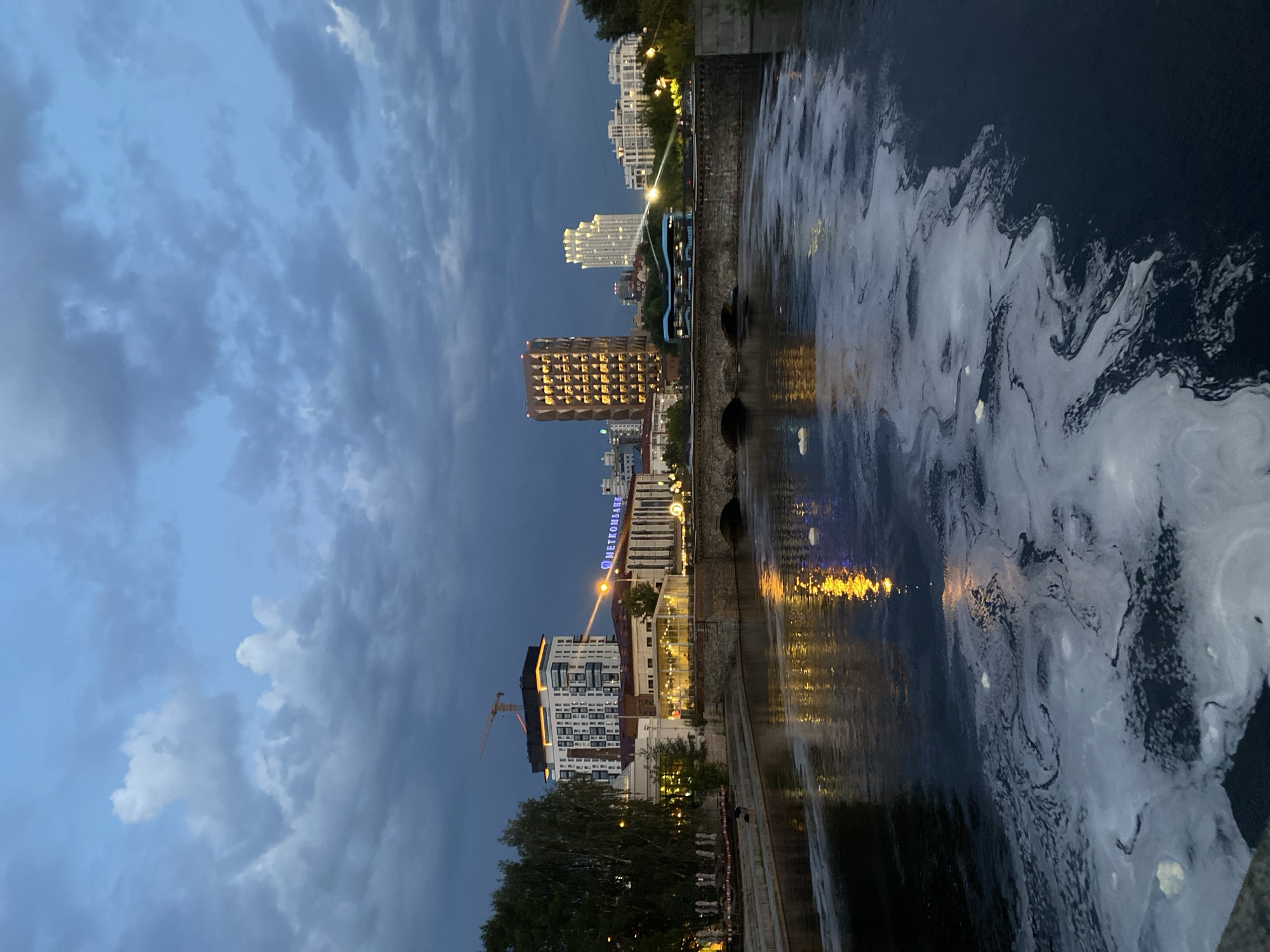 The image depicts the Iset River in Yekaterinburg, Russia, at dusk. The sky is a deep blue with dramatic clouds, hinting at recent or impending weather changes. The river surface is dark and reflects the warm yellow lights of the city's buildings and bridges, creating a shimmering effect. White foam, likely from natural causes or pollution, swirls on the water's surface, particularly in the foreground.
On the riverbank, several buildings are visible. The most prominent is a modern, white high-rise with orange accents at the top, featuring a construction crane on its roof. Adjacent to it is a building with large glass windows, illuminated from within. Further along the bank, the facade of a building with a sign reading "МЕТКОМБАНК" (Metcombank) is visible. Across the river, more buildings form the city skyline. A stone bridge with three arches spans the river. On the bridge, a double-decker bus is visible, suggesting it is a road bridge. A single person is faintly visible on the riverbank in the lower left portion of the image, appearing to be standing near the water.