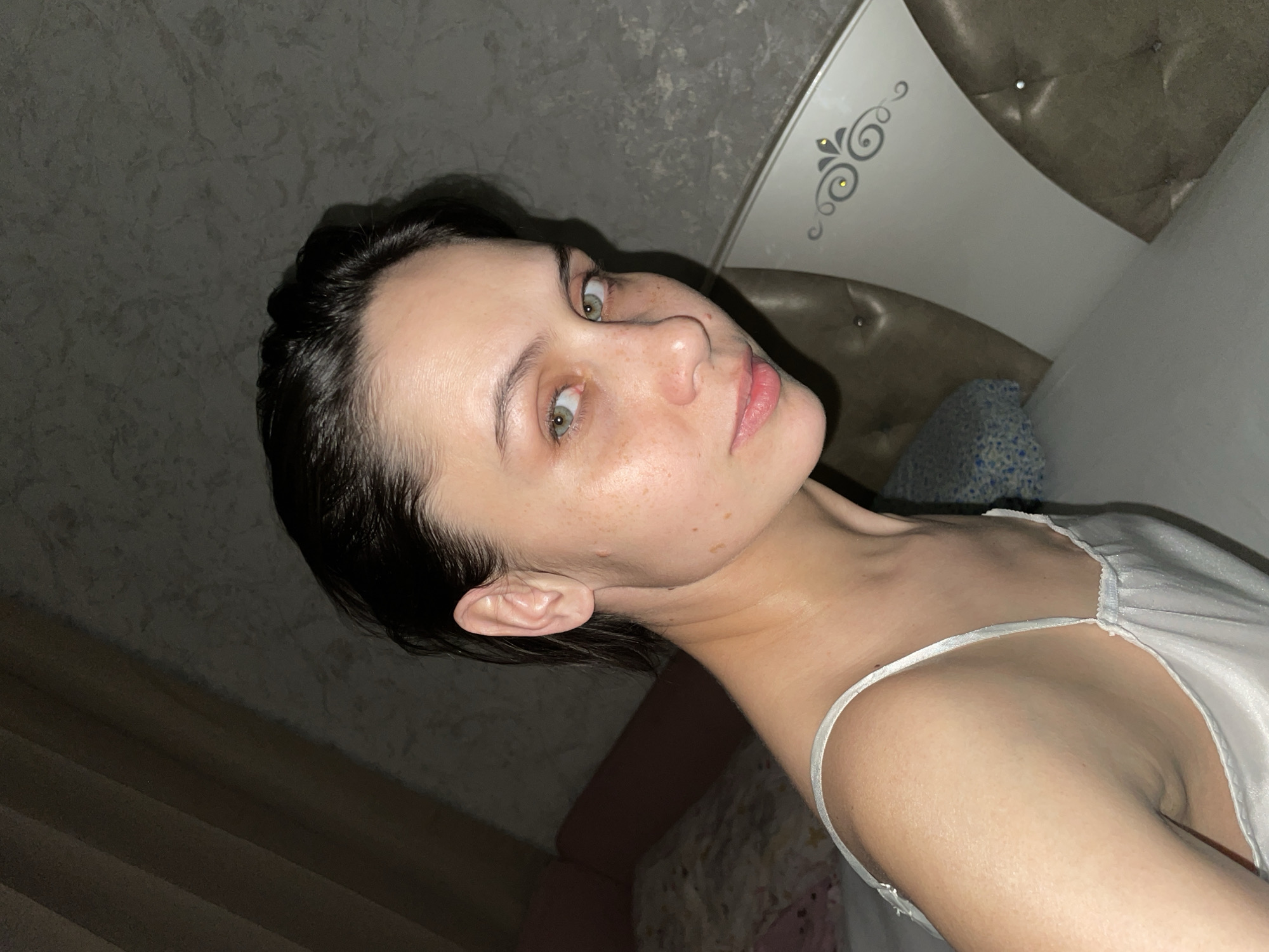 A young woman with fair skin and dark hair is lying in bed. She is wearing a white satin or silk top with thin straps. The background includes a headboard with decorative silver swirls and a gray patterned wall. The bed appears to be made with a white pillow and a blue patterned pillow. The lighting suggests an indoor environment, possibly a bedroom. There is no information to determine if the location is in the city of Izhevsk, Russia.