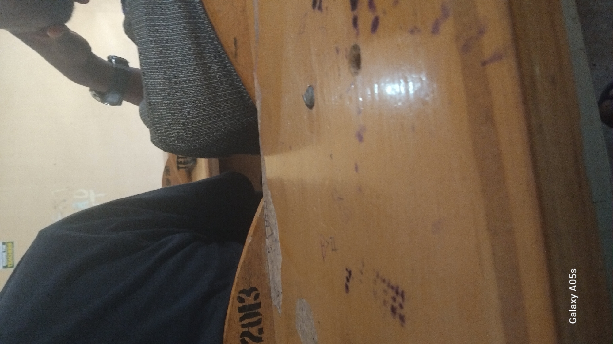 Two individuals are seated at wooden desks in a classroom. The desk in the foreground is a light brown, polished wood with visible scratches and markings. On the desk, there are several small, dark purple ink stains and a small, dark grey object, possibly a pebble, situated in a slight indentation. The back of the desk in the foreground shows the year "2013" stamped on its surface. The individual on the left is wearing a dark blue long-sleeved shirt, and their arm and shoulder are visible. The individual on the right is wearing a grey, textured, long-sleeved shirt and a watch on their left wrist. The background consists of a light-colored wall with some faint writing on it. The scene is lit by overhead lighting, creating reflections on the polished wooden surfaces. The image is identified as being taken in Yali, Nigeria, as indicated by the "Galaxy A05s" watermark, which is a mobile phone model.