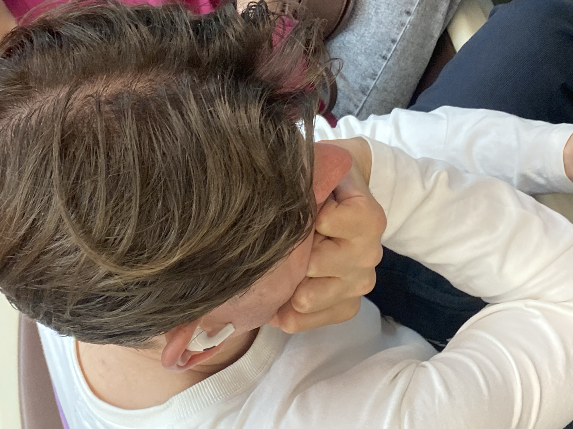 The image shows a close-up, overhead view of a person's head and upper body, resting their cheek on their hand. They are wearing a white long-sleeved shirt and navy blue pants. A white earbud is visible in their left ear. The person has medium-length brown hair with visible scalp. The background is blurred, showing hints of other people and furniture, suggesting an indoor setting. There is no information in the image to indicate the location in Moscow, Russia.