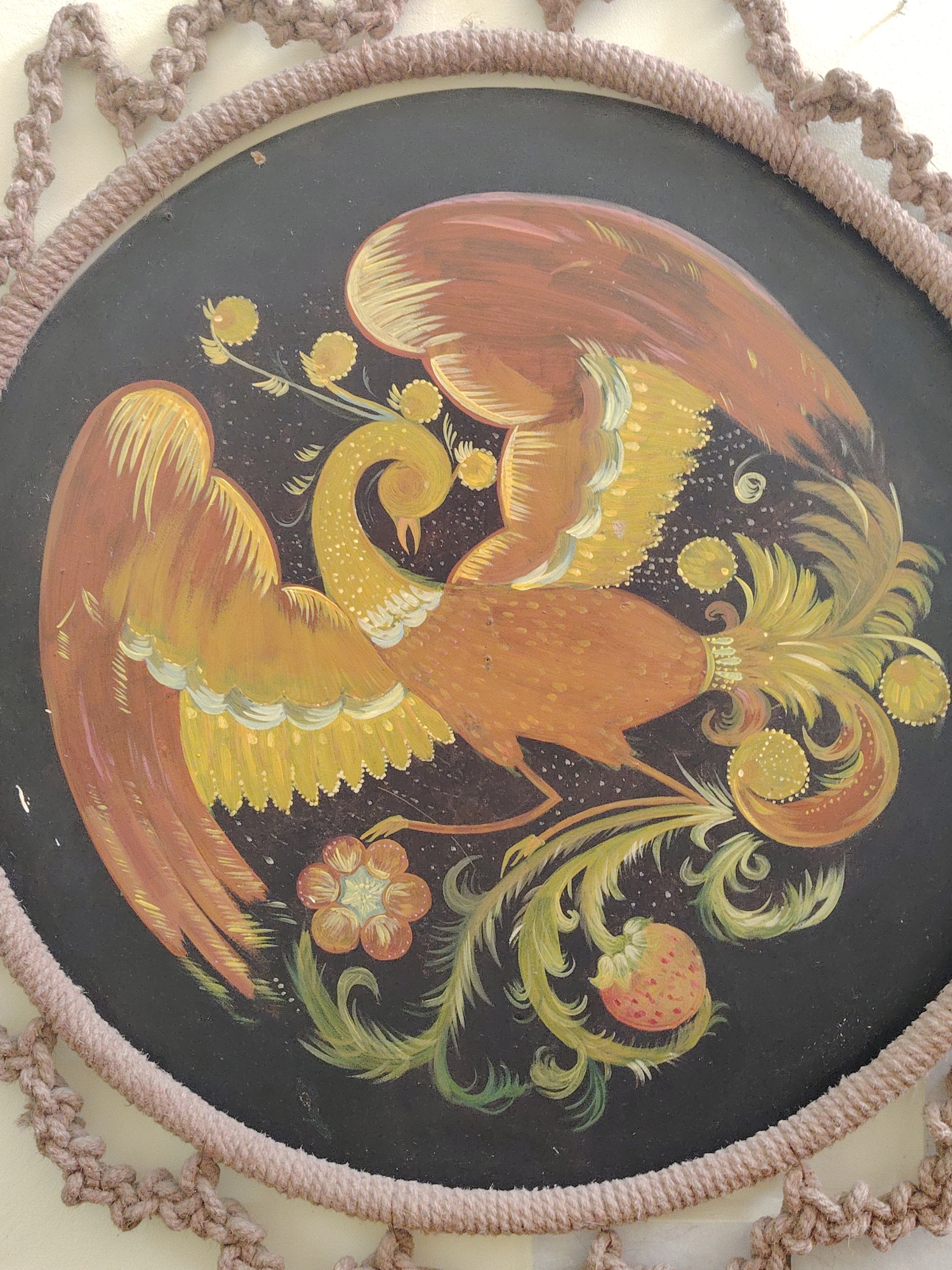 A close-up captures a round, dark lacquered tray or plate with a vibrant, folk-art style painting. The central motif is a stylized bird, reminiscent of a phoenix, with swirling golden and orange plumage. Floral and fruit elements, including a strawberry and flower, complement the bird's form. The tray is framed by a woven or crocheted border. This is likely a Khokhloma painting, a traditional Russian craft often featuring stylized birds and flora. Sarapul, Russia, is known for similar folk art traditions.