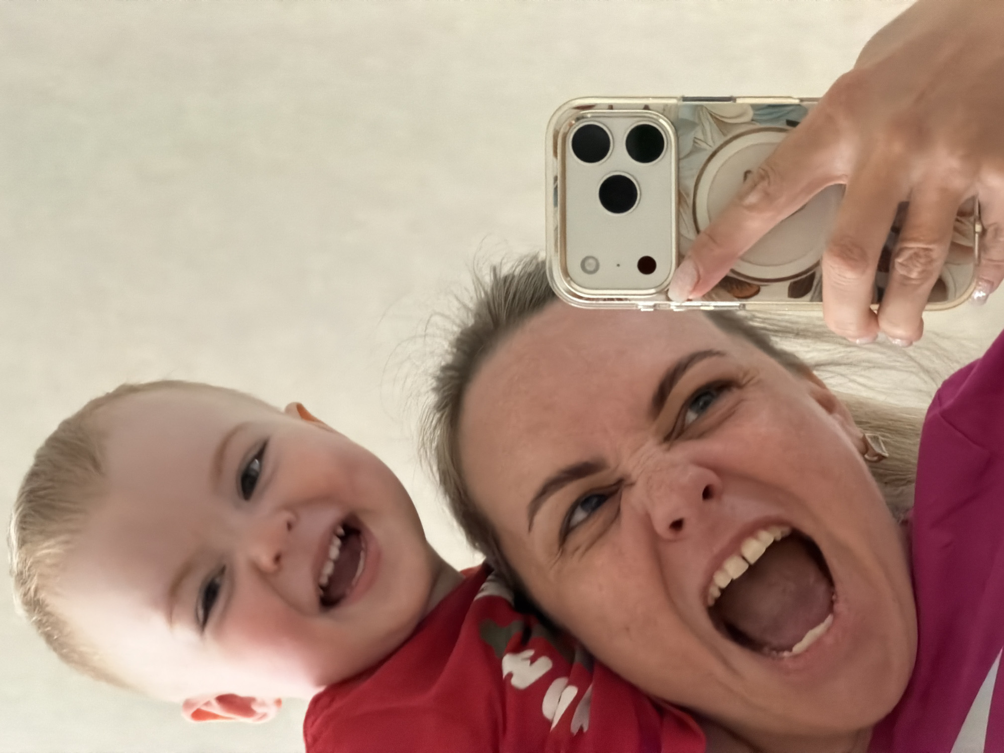 A woman and a young child, likely a mother and baby, are captured in a close-up, low-angle selfie, creating a dynamic and joyful composition. The perspective suggests the subjects are leaning over a smartphone placed on a flat surface, looking directly into the camera.
The woman, positioned on the right, has light hair and blue eyes, and is making a dramatically playful face with her mouth wide open, eyes squinted, resembling a humorous roar or scream. She is wearing a bright pink garment. Her left hand is visible at the top, holding a modern smartphone, which appears to be an iPhone with a triple-lens camera system, encased in a clear cover adorned with a subtle floral pattern and featuring a circular MagSafe attachment on the back.
The young child, on the left, has light hair and is wearing a red shirt. The child is smiling widely, mouth open in laughter, revealing baby teeth, and looking towards the camera with evident happiness.
The background is uniformly light and plain, suggesting an indoor setting. The overall mood is one of lighthearted fun, playfulness, and affection between the two individuals. No discernible text is visible on any objects in the image. This scene takes place in Votkinsk, Russia.