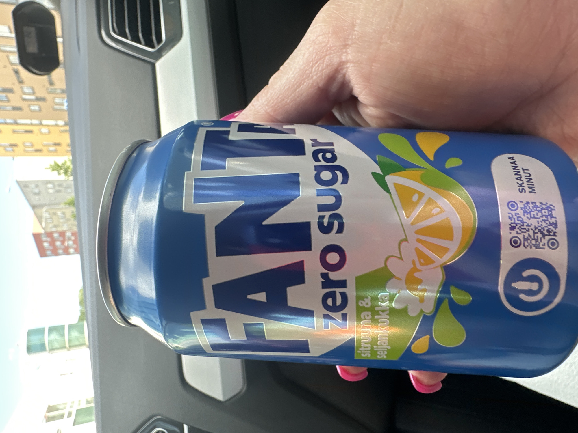A close-up, eye-level, outdoor shot shows a person's hand holding a blue Fanta Zero Sugar can with a lemon and elderflower flavor. The can features the Fanta logo in large, white, stylized letters, and the text "zero sugar." Below this, the flavor is listed as "sitruuna & seljankukka" in white text on a green background, accompanied by illustrations of lemon slices and flowers. A power button icon and a QR code are also visible on the can. The background suggests an urban environment with buildings and trees visible through a car window. The hand holding the can has bright pink fingernails, and the skin appears textured. The overall lighting indicates a sunny day.