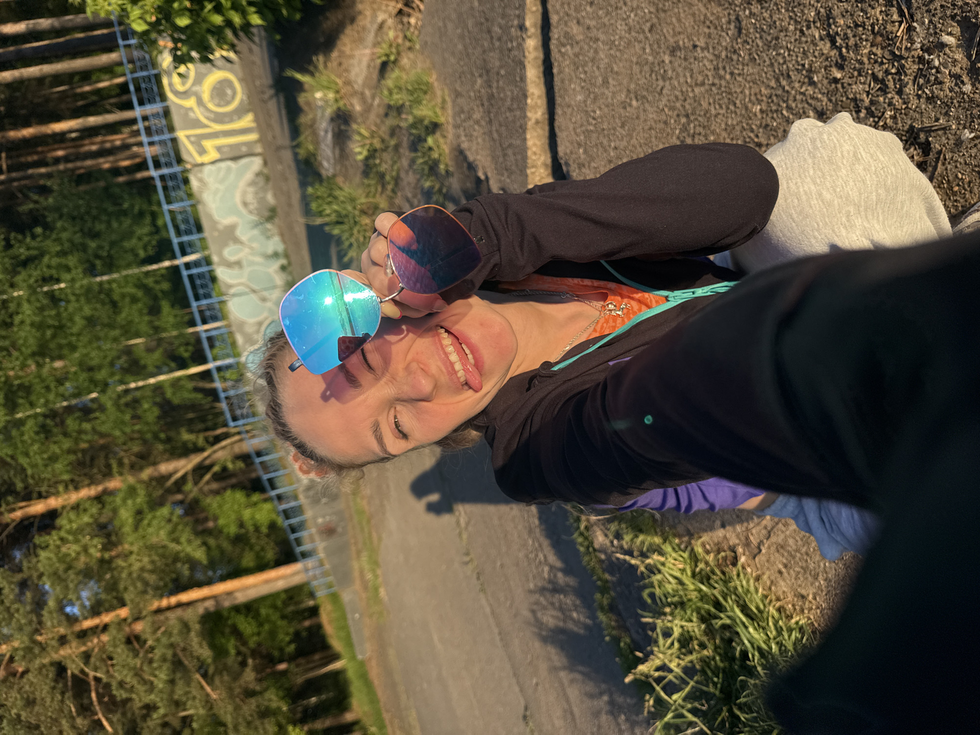 A playful selfie captures a young woman outdoors in Chaikovsky, Russia, during what appears to be golden hour or late afternoon. The warm, golden light casts soft shadows and illuminates the scene. The woman, possibly taking the photo herself, is positioned slightly angled, with her head tilted and her tongue playfully sticking out, indicating a cheerful and lighthearted mood. She is wearing a black long-sleeved top or jacket over a brightly colored, possibly orange and teal, inner layer, and light grey bottom wear.
She holds two pairs of sunglasses: one pair with reflective blue lenses is held up close to her face, reflecting the bright sky and surrounding greenery; another pair with orange-tinted lenses is held in her other hand. Her light-colored hair is visible around her face.
The background reveals a mix of natural and urban elements. To her left, there's a tall, dark metal fence or ladder-like structure, beyond which dense green trees are visible, suggesting a forest or park area. A concrete wall runs horizontally behind her, featuring yellow and light blue graffiti that includes what looks like the number "30" or "SO" in yellow. The ground appears to be a paved or gravel path, with patches of green grass and weeds growing along its edges. The overall impression is one of a casual, fun moment captured outdoors.