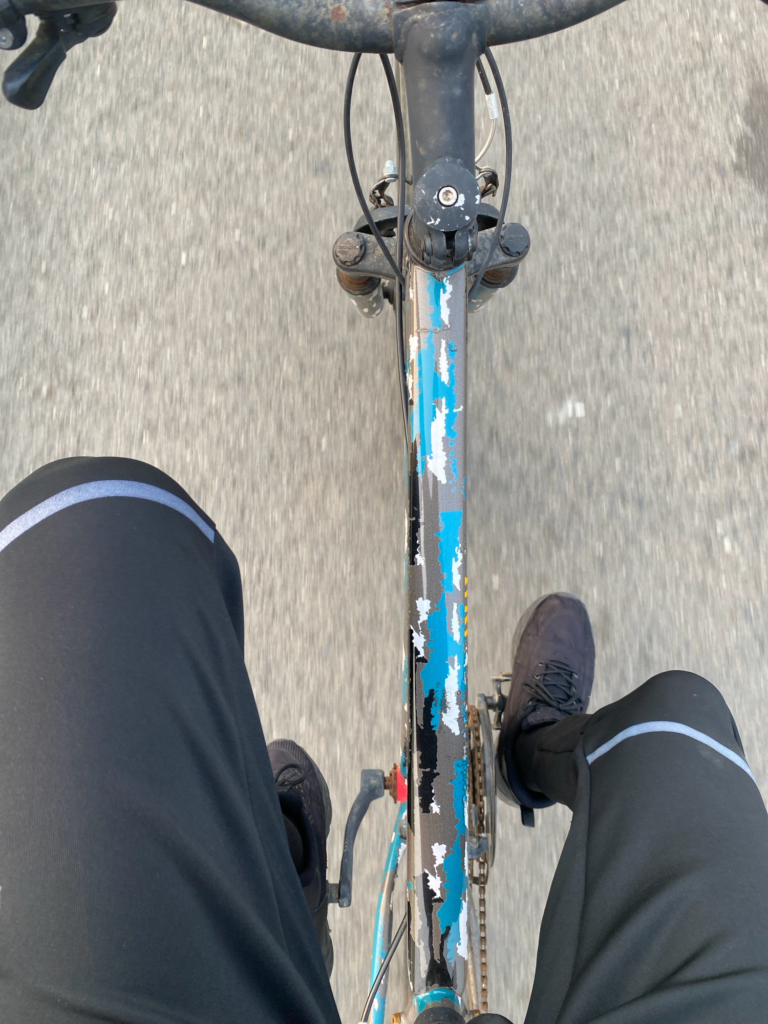 The image shows a first-person perspective of someone riding a bicycle on a paved road in Tanjung Pinang, Indonesia. The road surface appears to be asphalt, with some texture and faint white markings. The bicycle's frame is visible, with a worn, multi-colored paint job featuring blue, white, and gray patches. The handlebars, brake levers, and a portion of the front fork are also visible. The rider's black pants, which have a reflective stripe around the thigh area, and black athletic shoes are in the foreground. The motion blur of the image suggests the bicycle is in motion. The environment appears to be outdoors, with no other people or vehicles clearly discernible in the frame. The lighting suggests it could be daytime, possibly late afternoon due to the soft light.