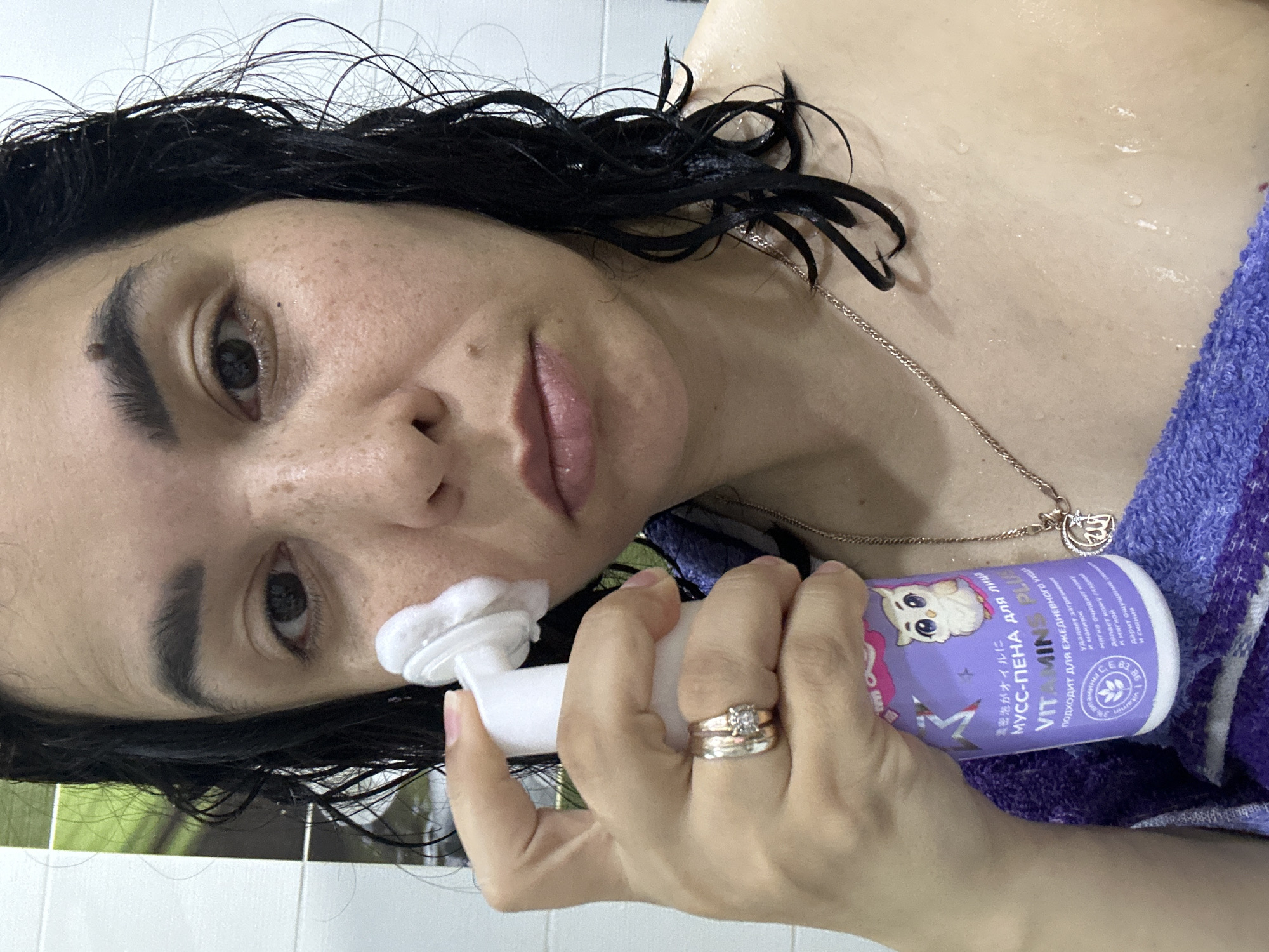 The image captures a woman in a bathroom setting, in the city of Unknown, Unknown. Her dark, wet, and curly hair is visible, framing her face. She has freckles on her skin. In her left hand, she holds a purple bottle with a white pump dispenser, from which she is applying a white foamy substance to the right side of her face, near her temple and cheekbone. The bottle's label features a cartoon owl and text in Cyrillic, identifying the product as a "Foam-Mousse for Face, VITAMINS PLUS," designed for daily care and makeup.
On her left ring finger, she wears a stack of three rings: two plain bands (one gold-toned, one silver-toned) and one silver-toned band with a single clear, square-cut stone. Around her neck, a gold-colored chain is visible, from which hangs a crescent moon pendant containing Arabic script. Her skin and hair appear wet, and water droplets are discernible on her bare shoulder and chest area. A purple towel is draped over her left shoulder and chest. The background consists of white and light green tiled walls.