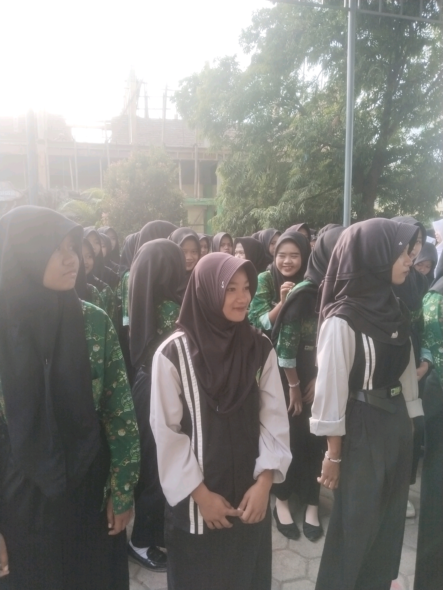 In the city of Pakulaut, Indonesia, a group of young women, likely students, are gathered outdoors. They are wearing hijabs and uniforms that include green tops with intricate patterns, and some are also wearing white shirts with black accents. The setting appears to be a school campus, with a building under construction visible in the background, along with trees and foliage. The lighting suggests it is daytime, possibly early morning or late afternoon due to the bright, hazy conditions. The women are standing close together, with some smiling and looking towards the camera or at each other. The ground is paved with light-colored tiles.