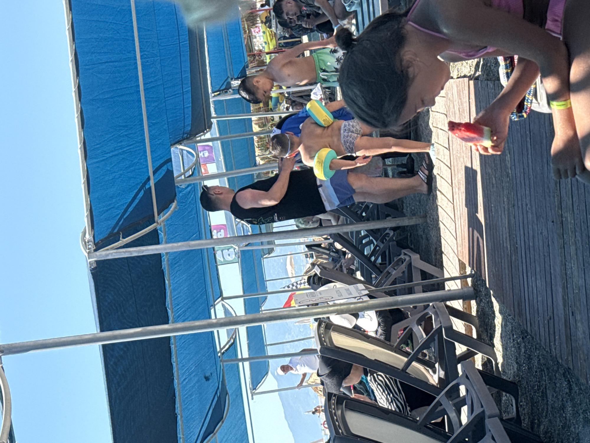 A photo of a beach with a large blue awning over a deck with lounge chairs. There are several people of various ages, including children, on the deck and beach. One girl in the foreground is eating watermelon. Two boys wearing inflatable armbands are on the deck. Another older boy in green shorts is walking. A man in a black shirt and blue shorts is talking with them. Other people are sitting and relaxing in the lounge chairs. In the background, the ocean, beach, and mountains are visible.