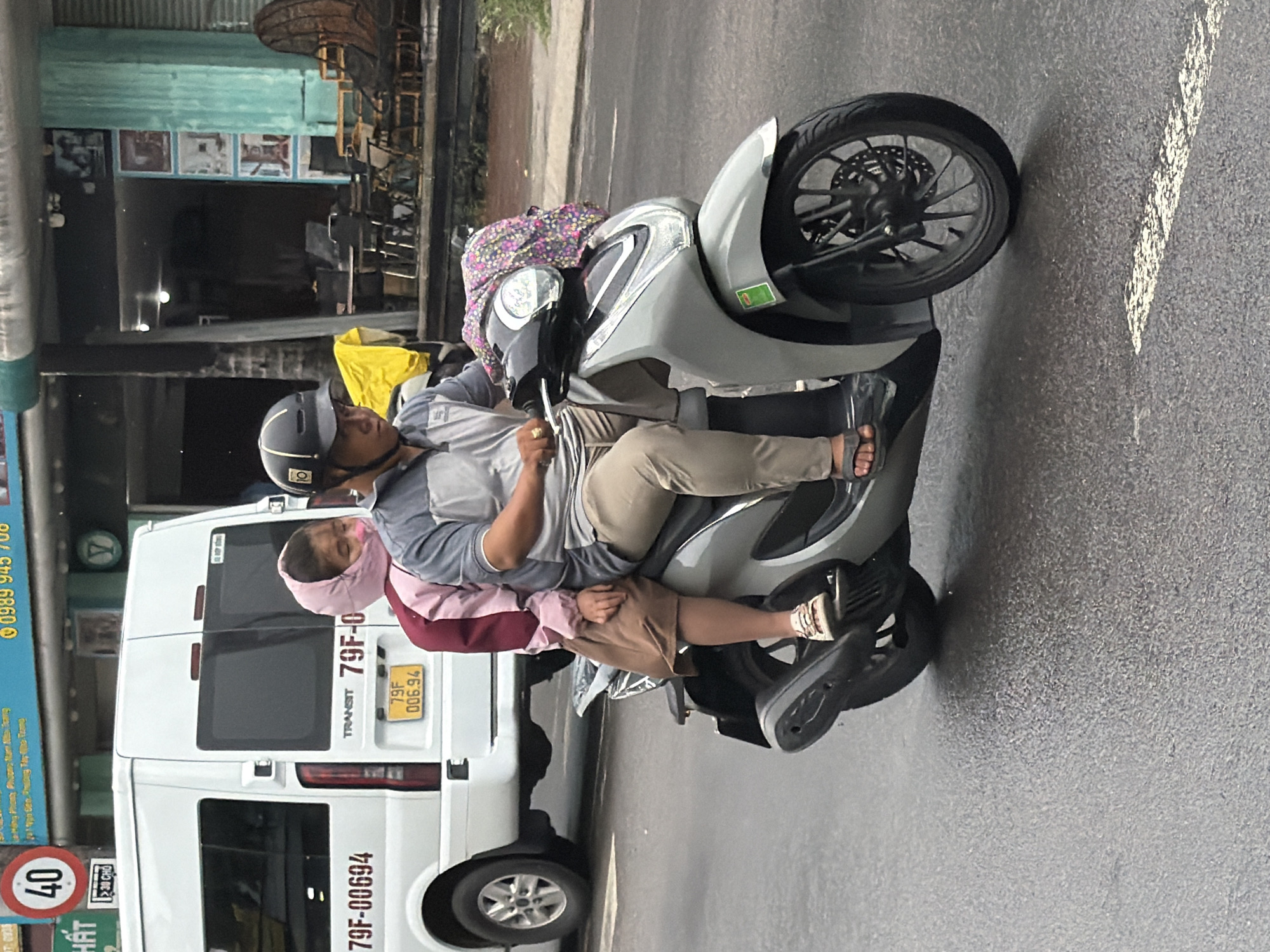 A man and a young child are riding on a gray scooter on a wet asphalt road in Nha Trang, Vietnam. The man, wearing a helmet and a gray polo shirt, is driving, while the child, wearing a pink headscarf and a brown skirt, is seated behind him. A white van with the license plate "79F-00694" is visible in the background, along with a speed limit sign indicating "40." Buildings with various signs and facades line the street. The image appears to be taken during the daytime.