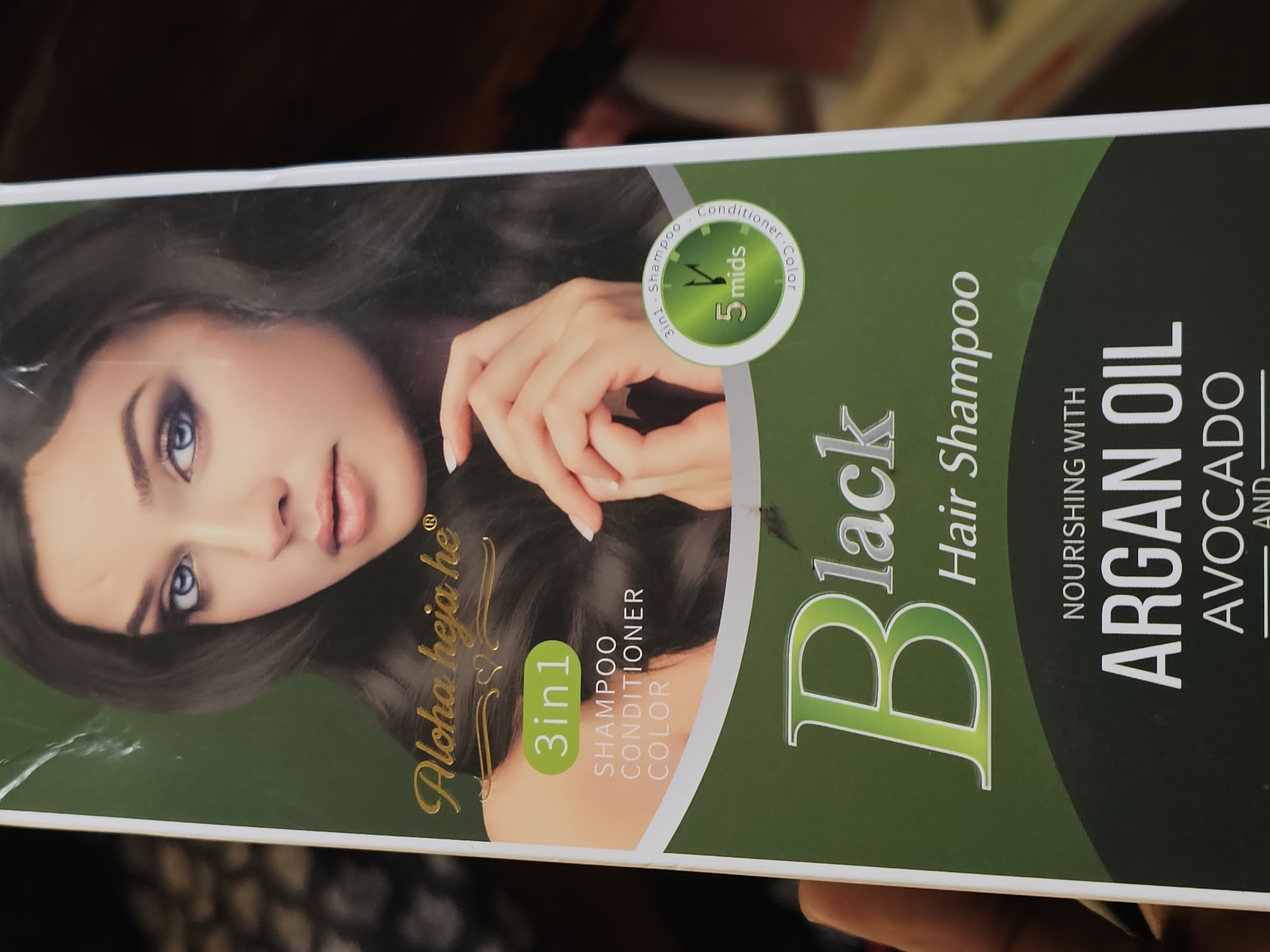 The image shows the front of a product box for "Aloha heja he" brand hair shampoo, specifically a "Black Hair Shampoo" variant. The box features a close-up image of a woman with dark hair and blue eyes, looking towards the viewer. The product claims to be "3in1" and is described as "SHAMPOO CONDITIONER COLOR". A circular graphic indicates "5 mids", suggesting a processing time. Below this, the product name "Black Hair Shampoo" is prominently displayed in large font. Further down, the text "NOURISHING WITH ARGAN OIL AVOCADO" is visible, indicating key ingredients.
The photograph is taken from a slightly elevated angle, with a hand holding the bottom left corner of the box visible. The background is dark and appears to be an indoor setting, possibly a retail environment or a home. There is no discernible information in the image that would allow for its specific location in Dar es Salaam, Tanzania.
