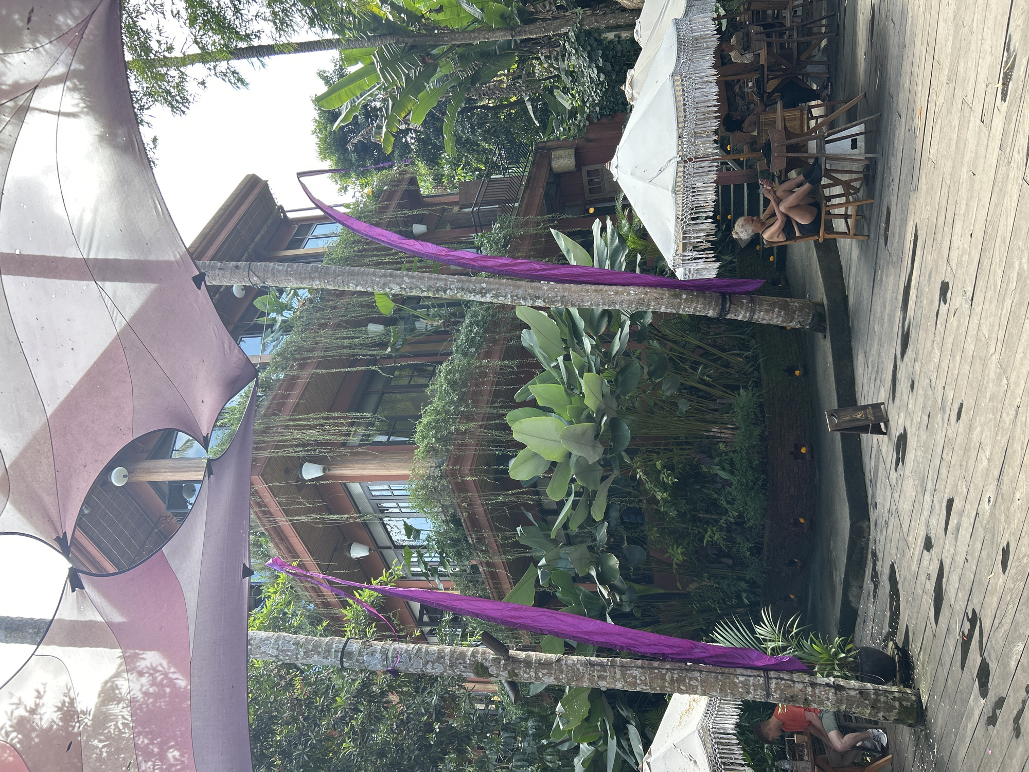 The image shows an outdoor seating area with a multi-story building in the background, likely in Ubud District, Indonesia. The building is covered in lush greenery and hanging plants, with wooden balconies and windows. Purple fabric streamers are draped from trees and around poles, adding a decorative element. White umbrellas with intricate fringed edges provide shade over some of the tables.
In the foreground, people are seated at tables. On the left, a person wearing an orange t-shirt and shorts sits at a wooden table. In the middle right, a woman with blonde hair sits on a chair, looking at her phone. Other people are visible at tables further back. The ground is made of wooden planks, with some wet patches suggesting recent rain. The overall atmosphere appears to be a relaxed outdoor dining or cafe setting surrounded by nature.