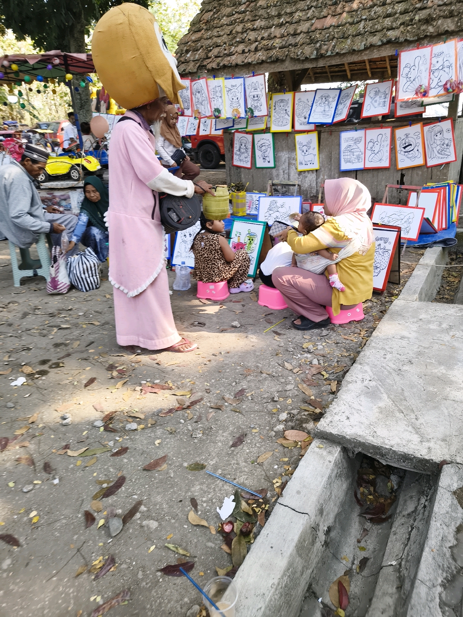 In Tlogorejo, Indonesia, a person in a large yellow duck costume stands near a stall displaying numerous framed coloring pages. To the right, a woman wearing a pink hijab and a yellow top sits on a pink stool, holding a baby. Another child in a leopard-print dress is seated on a similar pink stool to her left. In the background, several other individuals are visible, along with various items like bags, a yellow toy car, and a red vehicle. The scene appears to be an outdoor marketplace or event, with a dirt and leaf-covered ground and a structure with a tiled roof.