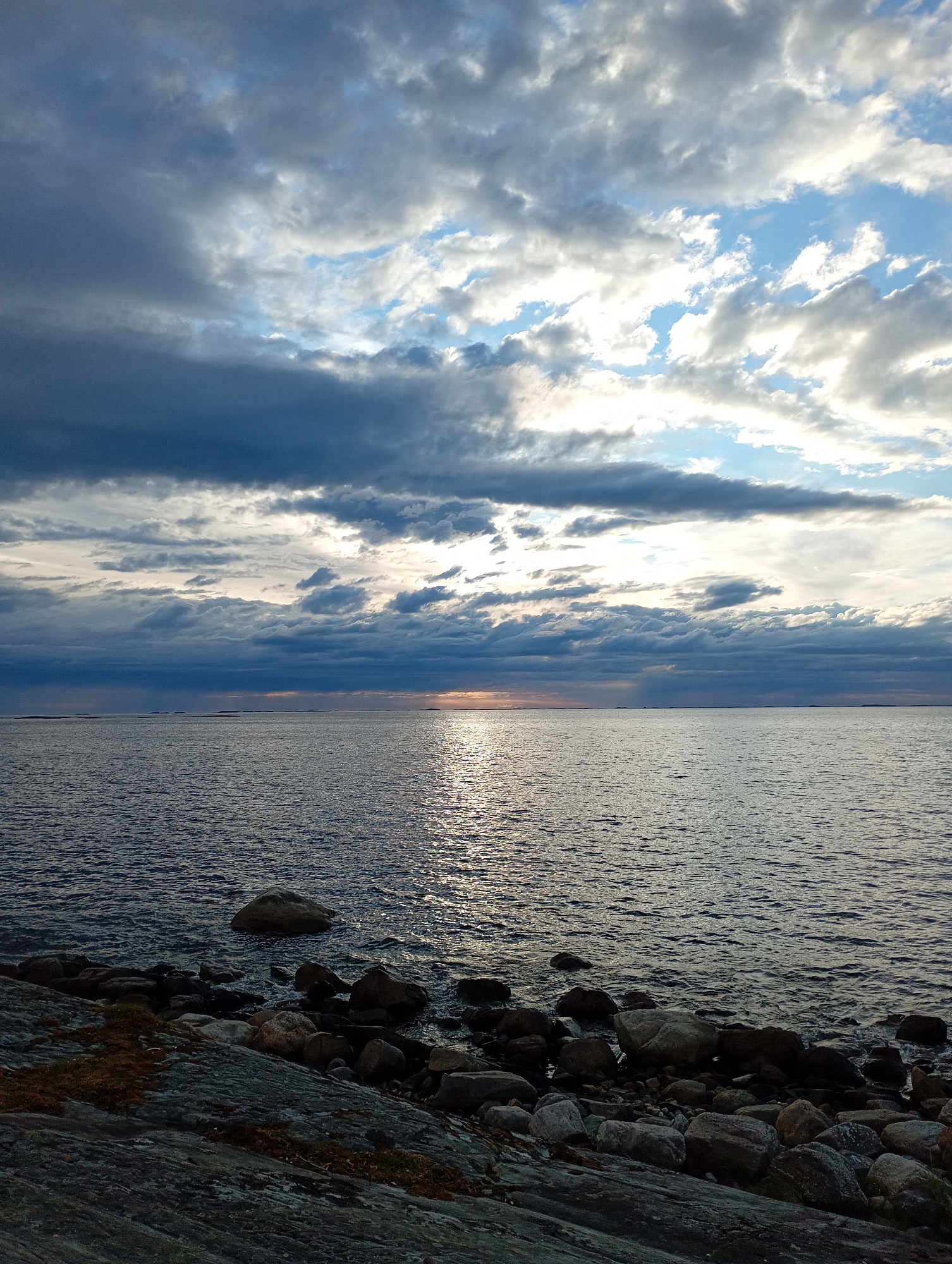 The image depicts a rocky shore on the left, meeting a calm body of water that extends to the horizon. The water reflects the light of a sun partially obscured by dramatic, cloud-filled skies. The clouds range from dark grey to bright white, with patches of blue sky visible. Several small islands or landmasses are faintly visible on the horizon. The foreground is dominated by various-sized grey and brown rocks, some with patches of dry grass or moss. The water is a deep blue-grey, with ripples catching the sunlight, creating a shimmering effect. The overall impression is of a serene, possibly overcast, coastal landscape. The location is unknown.