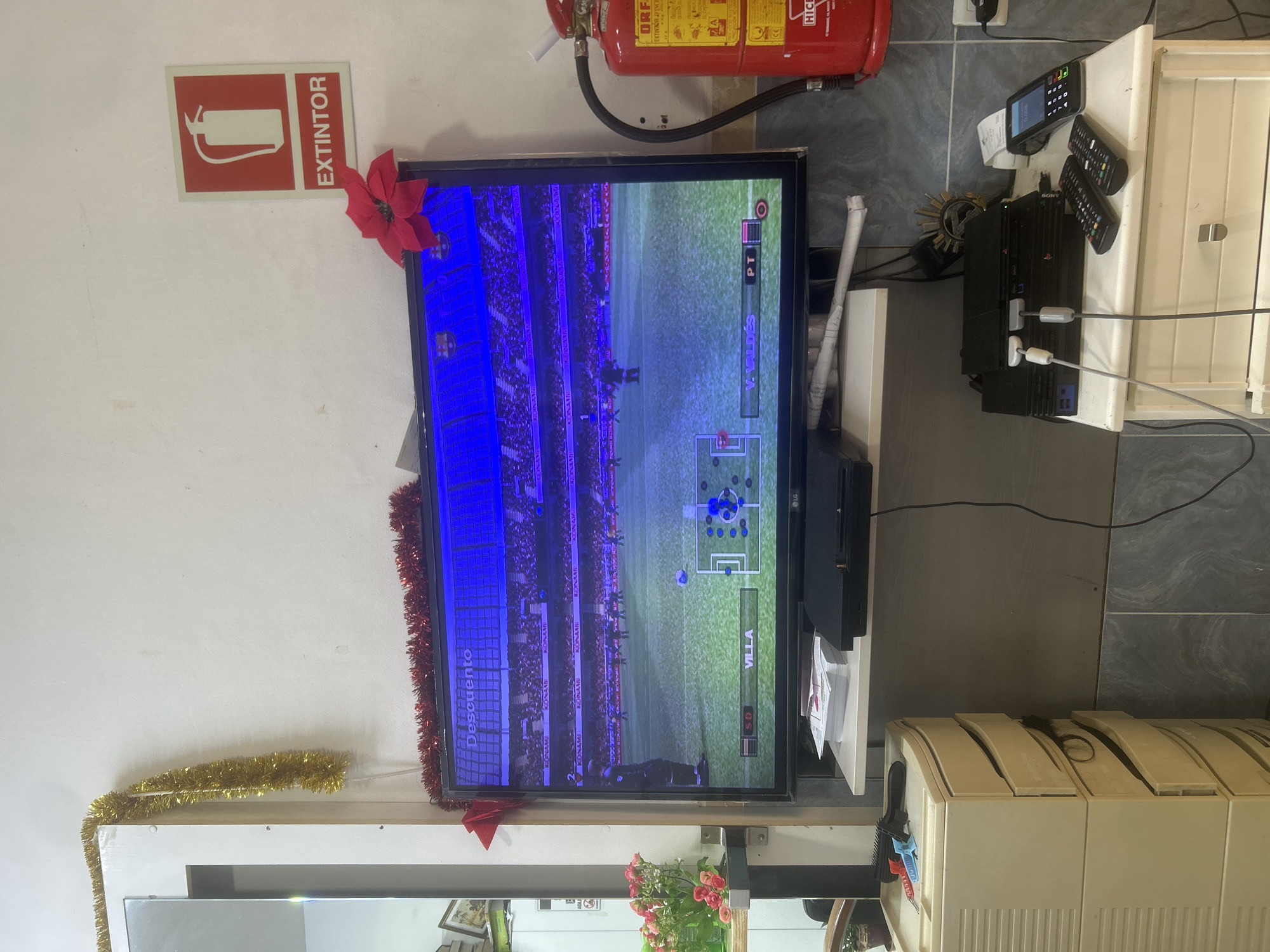 The image shows a cluttered interior space, possibly a shop or small business. A television screen displays a soccer video game with the name "Villa" visible on the screen. Christmas decorations, including tinsel and a red poinsettia, adorn the TV and surrounding area. Above the TV is a fire extinguisher and an "EXTINTOR" (Spanish for extinguisher) sign. To the right of the TV, on a white surface, there is a Sony PlayStation 2 console, a handheld device, and two remote controls. The flooring appears to be tiled. There is no visible indication to confirm this is in Barcelona.