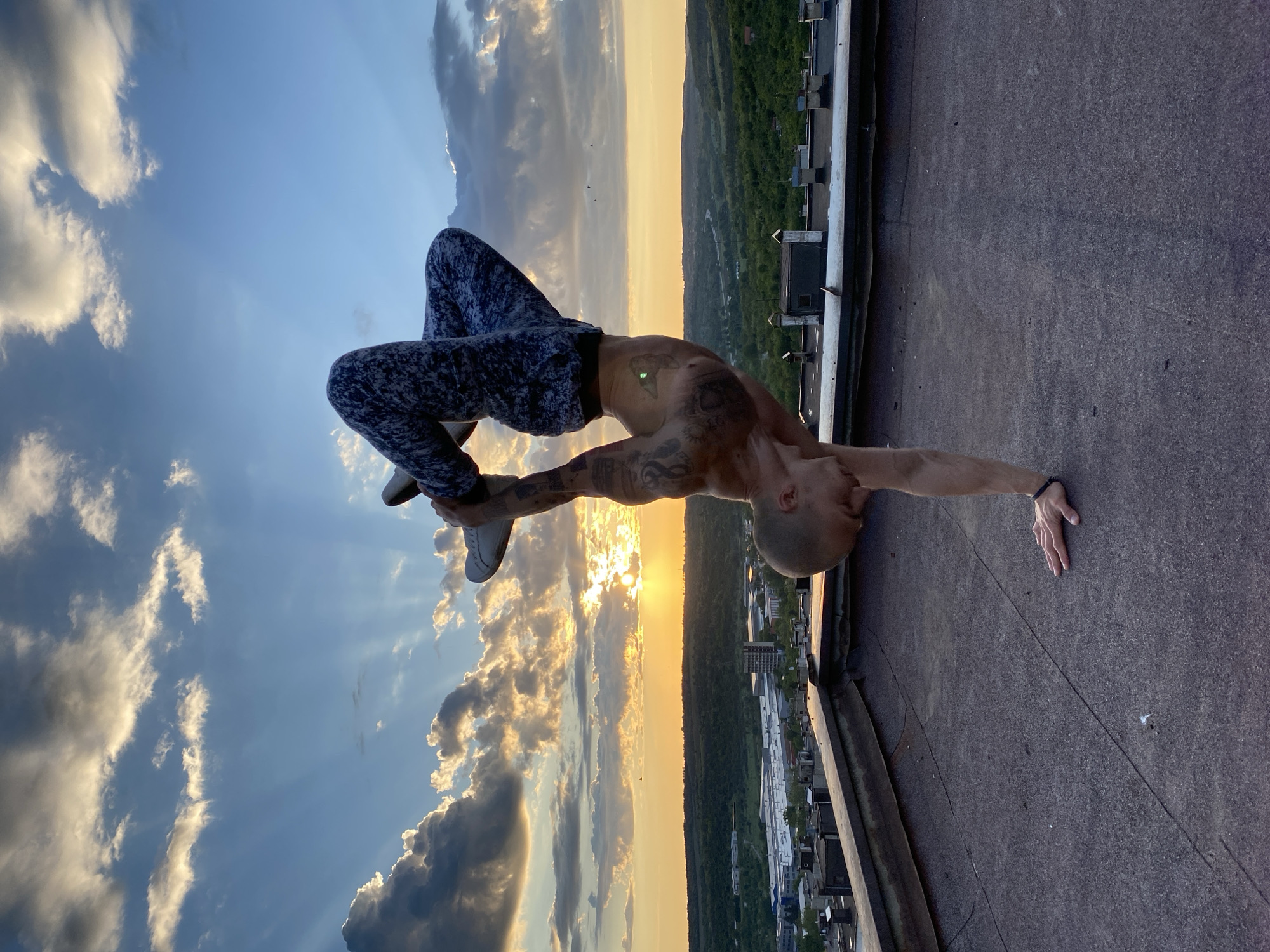 A man is posing on the edge of a rooftop in Pleven, Bulgaria. He is wearing patterned pants and sneakers. The sun is setting over the city in the background. The rooftop is flat and grey. The man's body is arched back, with one hand on the rooftop and his feet touching his other hand. He has tattoos on his arms and torso.