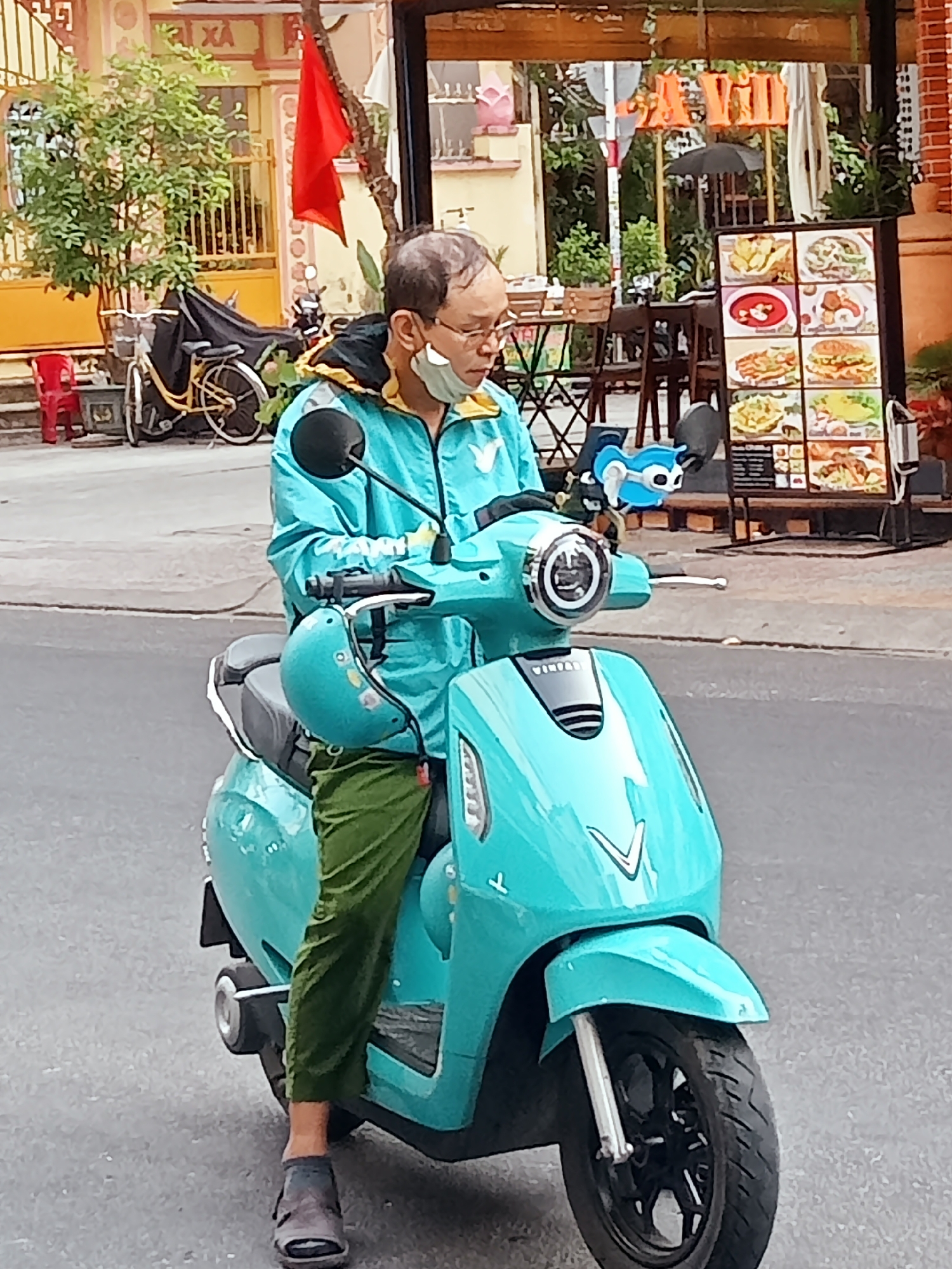 A man rides a turquoise scooter in Nha Trang, Vietnam. He wears green pants, a turquoise jacket, and a face mask. A matching helmet hangs from the scooter. A blue cartoon character is attached to the scooter's handlebar. In the background are buildings, trees, a red flag, a bicycle, and a restaurant menu board.