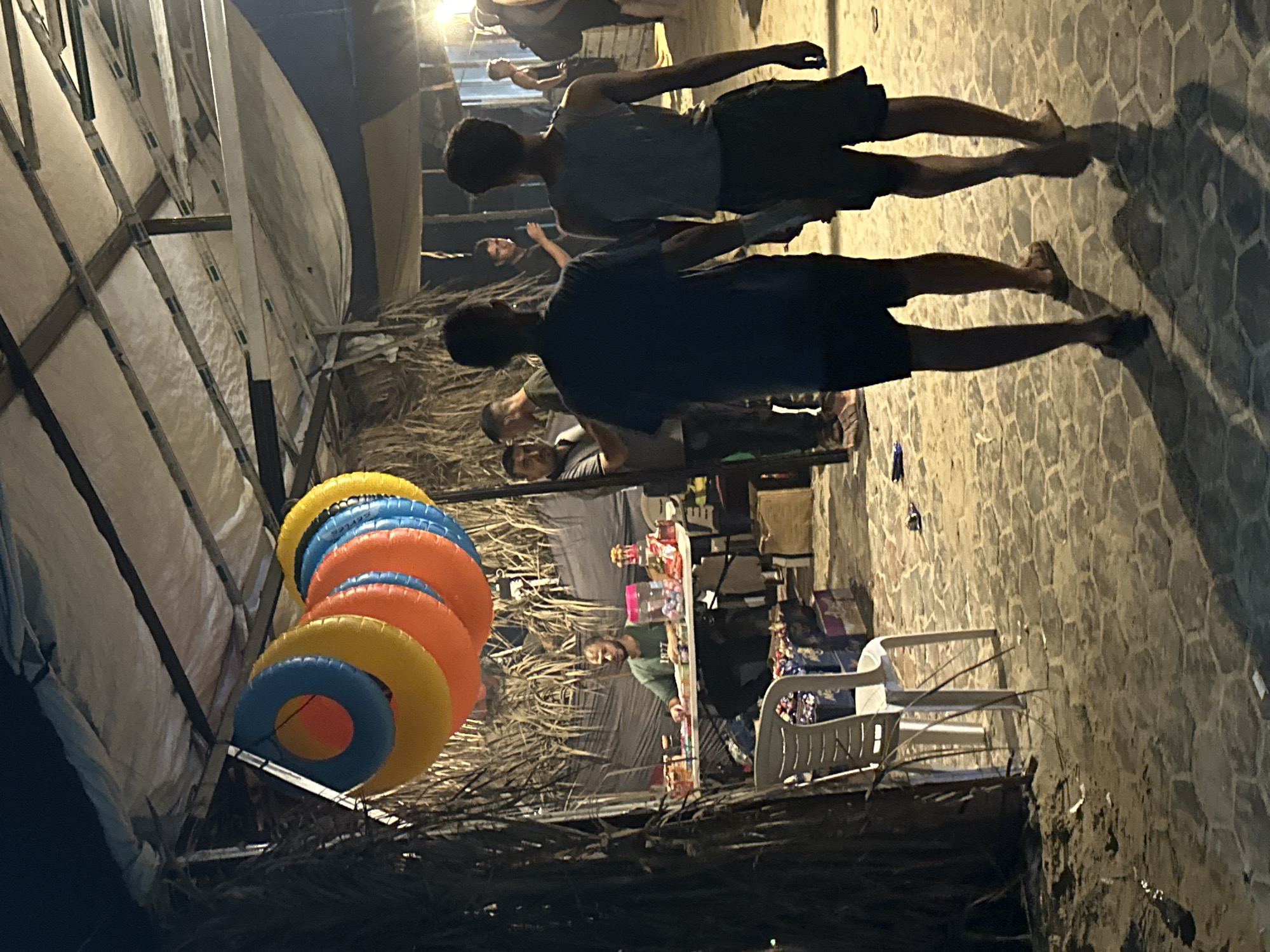 In the city of Gaza, an outdoor market scene unfolds at night. A man in a green t-shirt sits behind a table laden with various goods, possibly snacks or small items, some of which are in clear plastic containers. In the foreground, two young men walk away from the camera, their backs to the viewer, one in a dark blue t-shirt and the other in a light blue tank top and dark shorts. Behind them, other individuals are visible in the dimly lit background, suggesting a bustling atmosphere. A string of colorful inflatable rings hangs above the table, adding a playful element to the scene. The ground is paved with hexagonal stones, and palm fronds and straw structures create a rustic ambiance. The overall impression is of a lively evening market with a mix of commerce and casual social interaction.