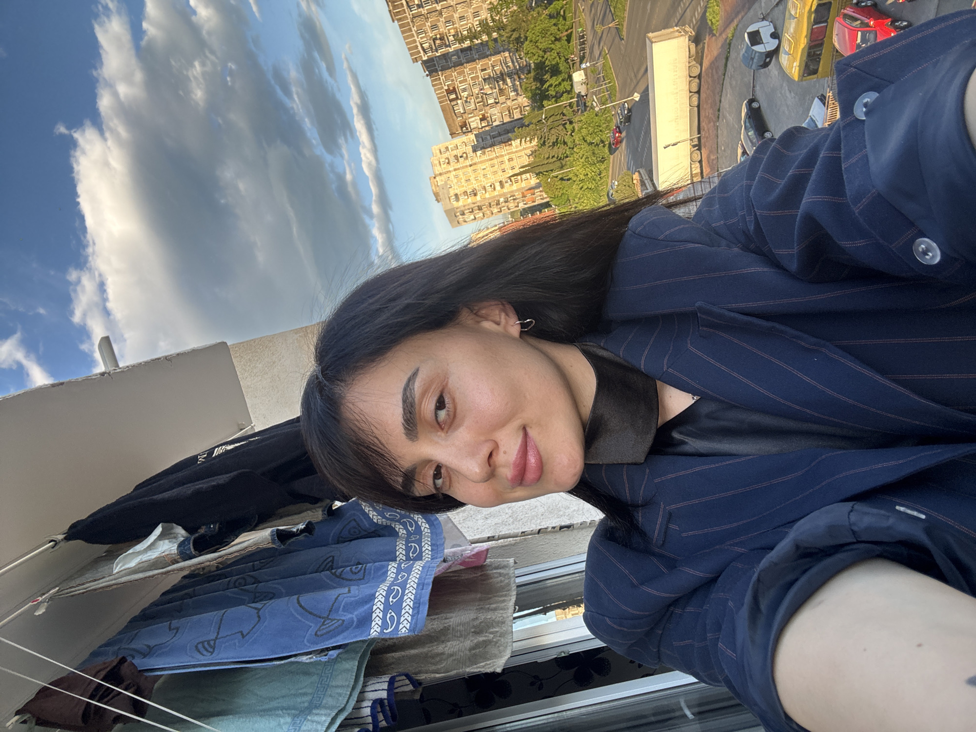 A medium-shot, angled selfie shows a person with dark hair, wearing a striped blue blazer over a black top. The person is looking slightly upward and smiling. Behind them, there are buildings, possibly apartments, and a street scene with cars and a white truck. In the lower left corner of the frame, laundry is hanging on a line. The city is Tbilisi, Georgia.