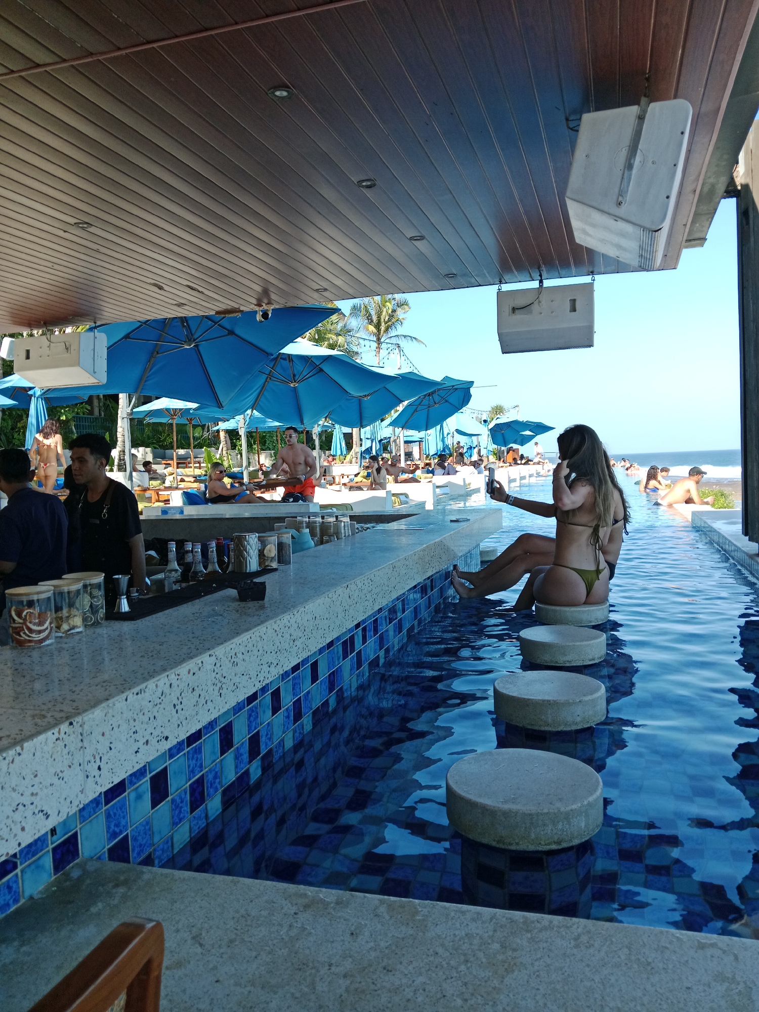 In Kuta Selatan, Indonesia, this image captures a lively scene at a poolside bar. The foreground features a white speckled bar counter adorned with jars of what appear to be dried fruit and other bar supplies. Behind the counter, a male bartender in a black apron is visible. To the right, the bar extends into a swimming pool where a woman in a bikini is seated on a circular stool, holding a smartphone and appearing to take a selfie. More people are seen relaxing by the pool, some under blue umbrellas, and in the background, the ocean and a palm tree are visible. The environment is bright and sunny, suggesting a vacation or leisure setting.