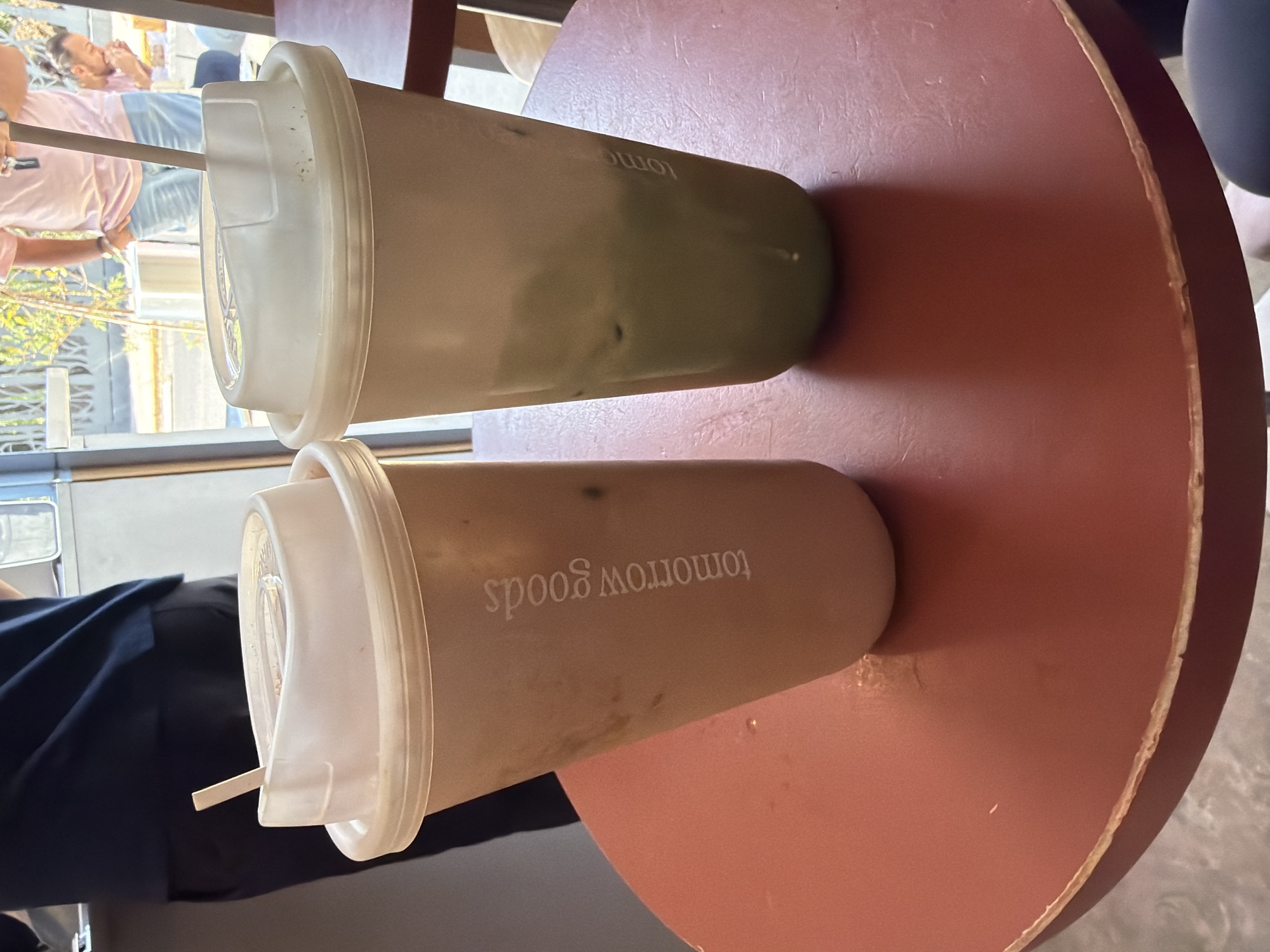 Two closed beverages are on a red round table. One beverage is green and the other is brown. Both have white lids and straws. The brown beverage has the words "tomorrow goods" written on it in reverse. People are visible in the background through a window. The scene is likely in Puebla, Mexico.