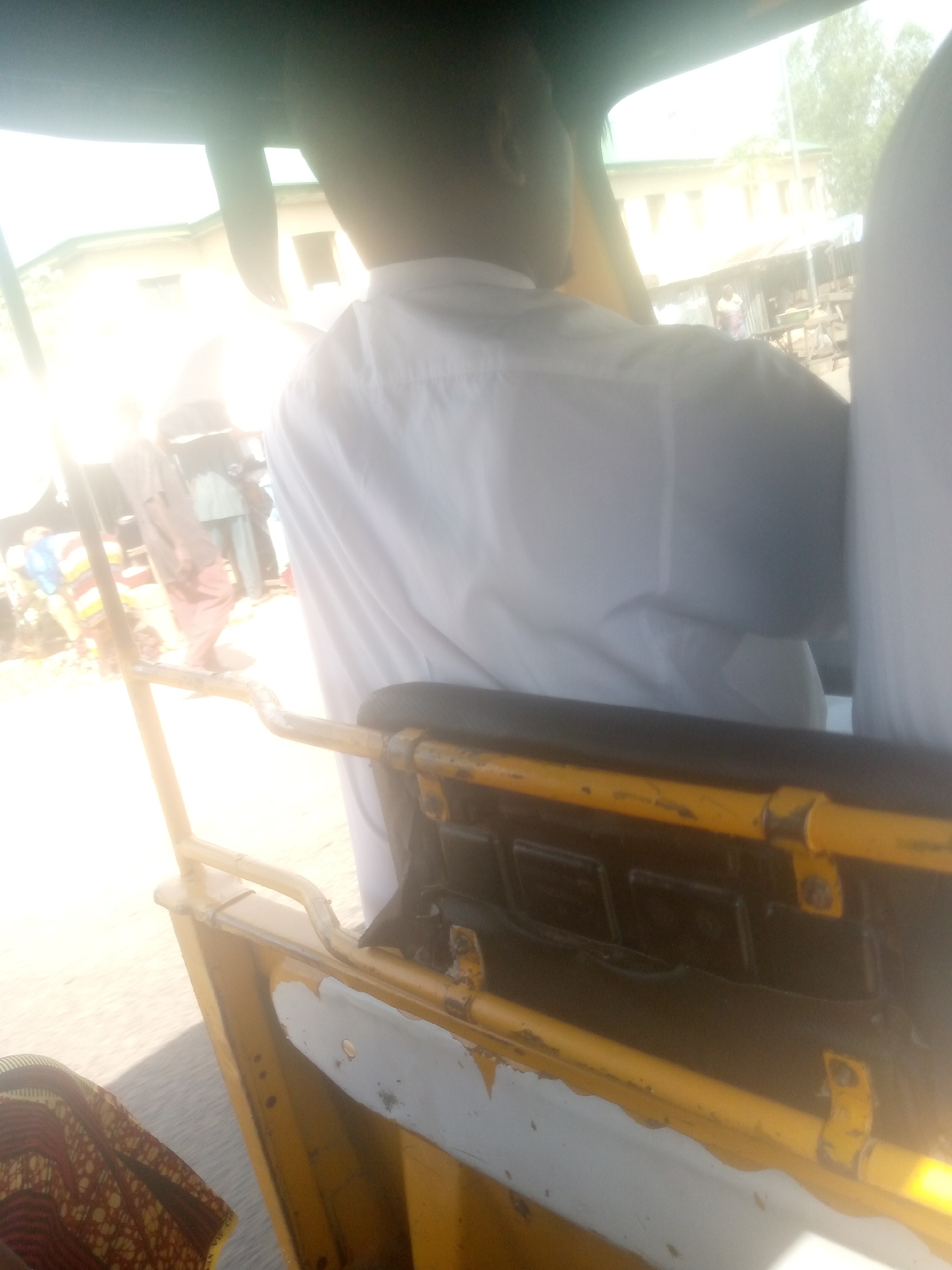 The image captures an interior view of a yellow auto-rickshaw, likely in Jalingo, Nigeria, as indicated by external contextual information. The perspective is from within the vehicle, looking towards the front passenger area and out through the windshield. A person wearing a white shirt is seated in the driver's seat, facing away from the camera, with their back and right shoulder visible. Sunlight is intensely bright, coming from the left, casting lens flares and washing out details of the environment outside. In the background, through the windshield, a building with multiple windows and a corrugated metal roof structure are partially visible. The yellow metal frame of the auto-rickshaw's passenger enclosure is prominent in the foreground, showing signs of wear and tear. A portion of a patterned fabric, possibly a garment or seat cover, is visible in the lower left corner. The scene appears to be set on a street or road, with some out-of-focus figures and structures suggesting an outdoor, possibly market or urban, setting. The overall impression is of a candid snapshot taken while the vehicle is in motion or stopped at a location with bright ambient light.