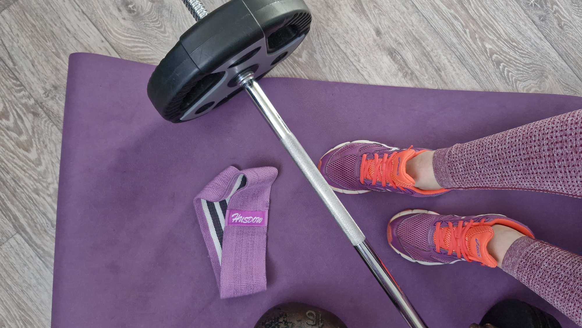 A top-down shot reveals a purple yoga mat on a light-colored wooden floor. A barbell with a black weight plate rests diagonally across the mat. Adjacent to the barbell, a folded purple resistance band with "Huisdow" printed on it is visible. Two purple running shoes with bright orange laces are positioned near the right edge of the mat, worn by a person whose lower legs, clad in purple textured leggings, are visible. The scene gives the impression of a home gym setup in a city of Unknown, Unknown.
