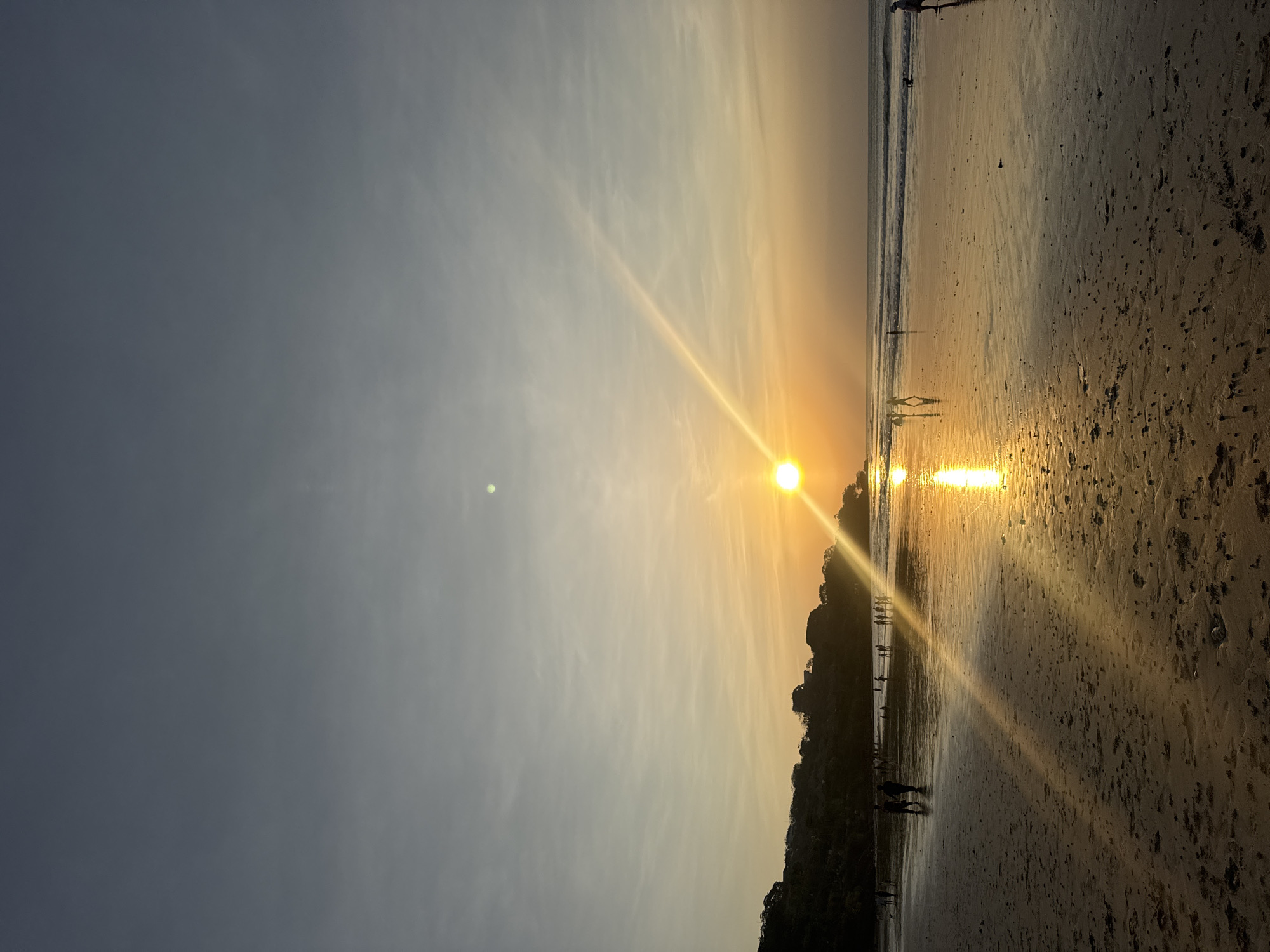 The image captures a serene sunset scene on a beach in Kuta Selatan, Indonesia. The sky transitions from a muted grey at the top to a warm gradient of yellow and orange as it nears the horizon. A bright, full sun is setting on the right side of the frame, casting a strong golden reflection on the wet sand and the shallow water. The foreground shows a sandy beach with footprints and scattered debris. A line of silhouetted figures, appearing to be people walking or standing, is visible along the water's edge, stretching from the left to the right of the frame. Some figures are closer to the viewer, while others are further away, creating a sense of depth. The ocean has gentle waves rolling in. On the left side of the image, a dark, tree-covered hillside rises, with some structures or lights faintly visible on its crest. The overall atmosphere is tranquil, with the warm glow of the sunset dominating the scene.