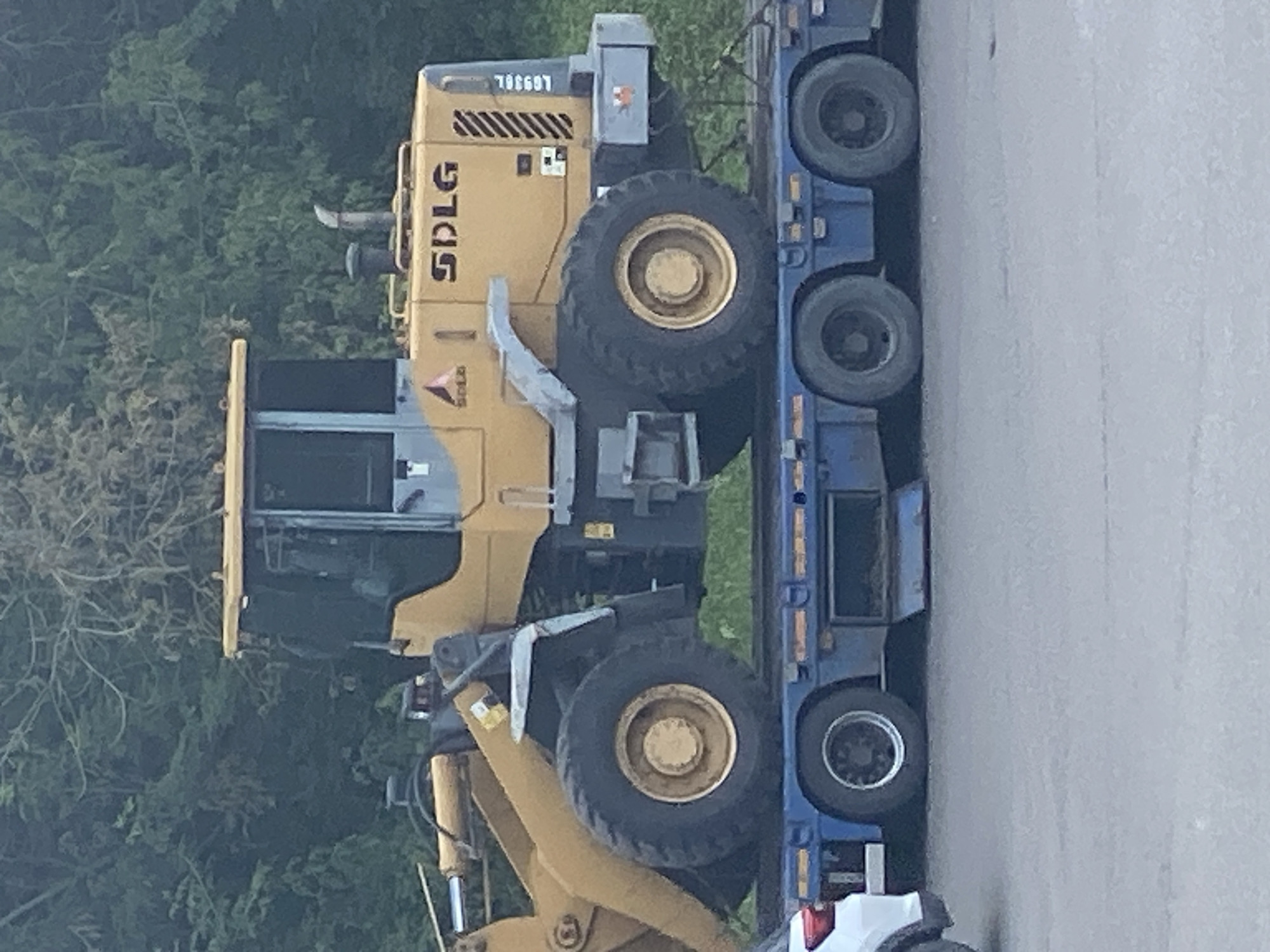 A yellow SDLG wheel loader, model LG950L, is loaded onto a blue flatbed trailer. The loader is positioned facing right, with its bucket lowered. The trailer has dual axles with large black tires. The scene is outdoors, with a blurred background of green trees and foliage. The ground is paved, suggesting a road or industrial area. There are no people visible in the image. The lighting appears to be overcast, indicating a cloudy day. The image quality is moderate, with some graininess. The location is the city of Novokuznetsk, Russia.