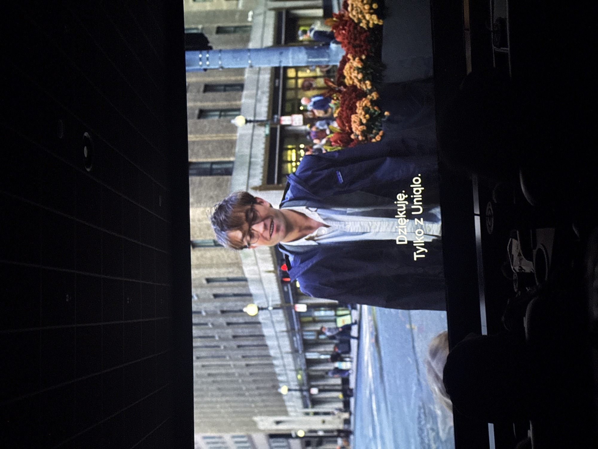 The image shows a movie screen displaying a scene with a man walking on a city street. The man is wearing a white shirt and a navy jacket. A subtitle in Polish reads "Dziękuję. Tylko z Uniqlo". The street appears to be in a North American city, with buildings, street lights, and people in the background. The presence of the Polish subtitle suggests the movie is being shown in Poland or to a Polish-speaking audience. It's impossible to pinpoint Warsaw, Poland as the specific location based on the visual elements of the scene displayed on the screen. The photograph was taken from a seat in the movie theater.