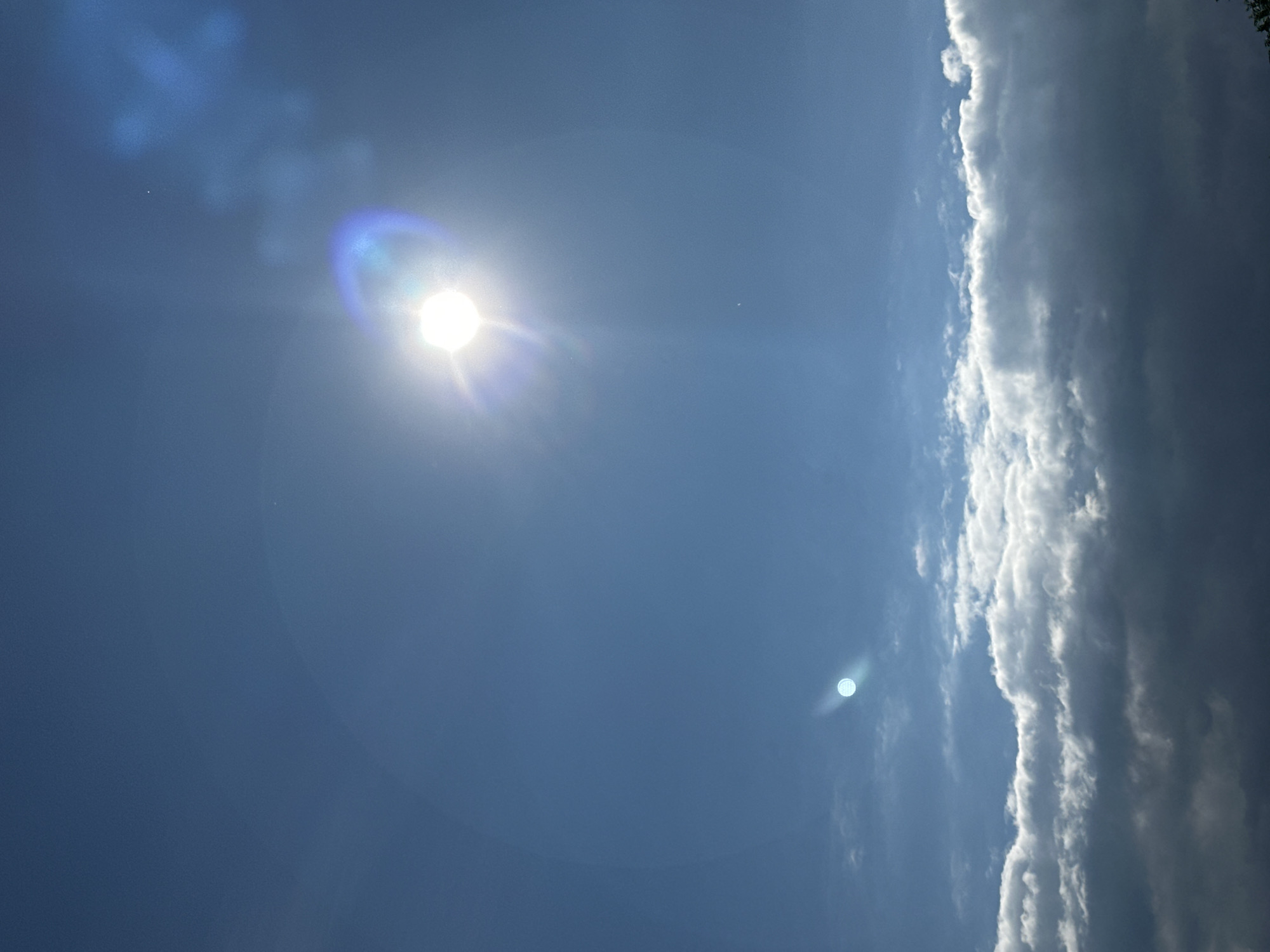 The image is a photograph of the sky, taken from an unknown location in an unknown city. The sun is visible, shining brightly and causing lens flare, with a ring of light and streaks of light emanating from it. There are some clouds in the sky; a few wispy clouds are higher up, and larger, darker clouds are closer to the bottom of the frame. The sky is a clear blue.