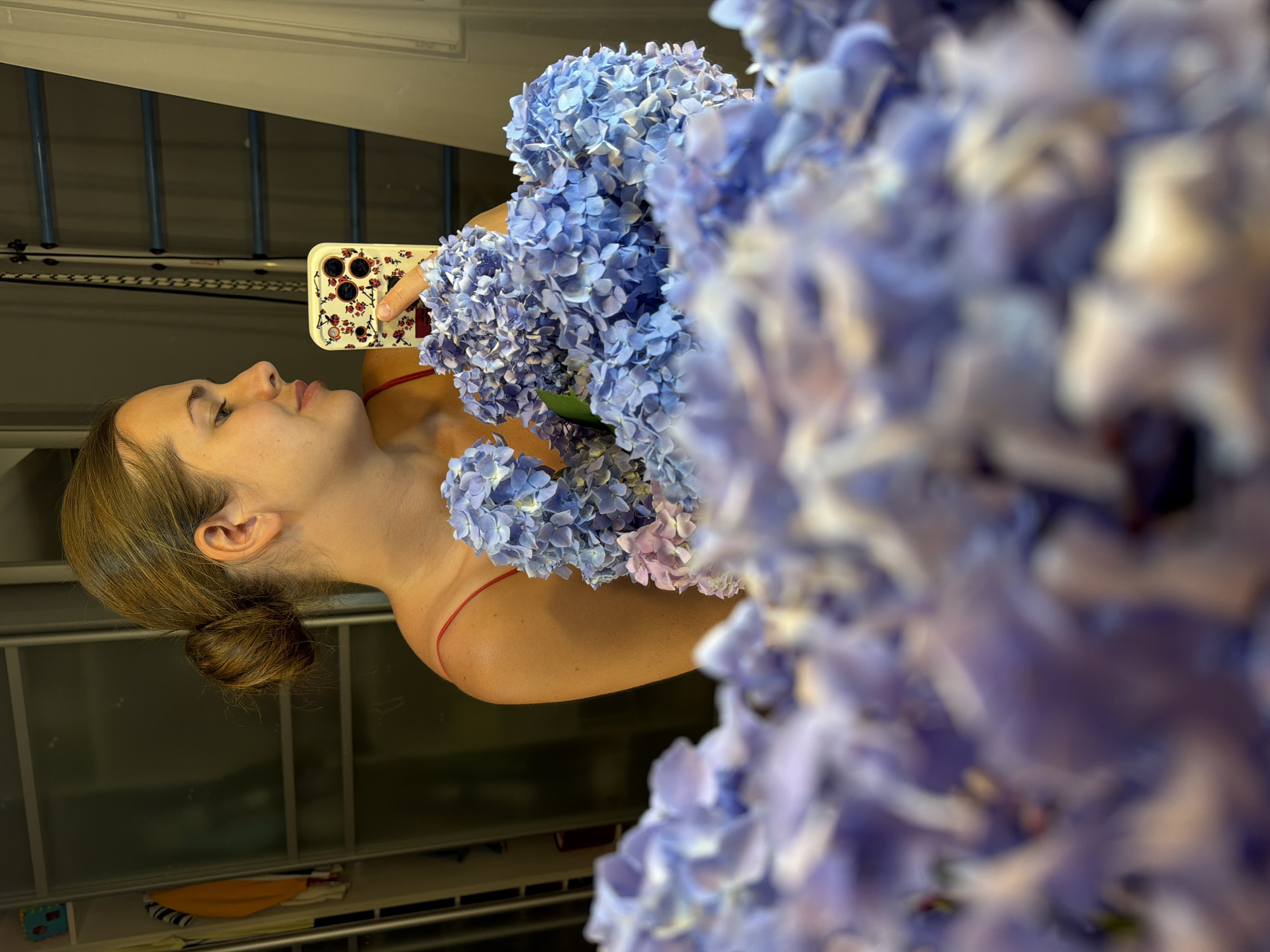 A woman with her hair in a bun is taking a selfie in a mirror. She is holding a smartphone in her right hand, and her left hand is holding a bouquet of blue hydrangeas. The hydrangeas are in the foreground and appear to be out of focus, while the woman and the mirror are in focus. The woman is wearing a red camisole. In the background, there are closets, and a ladder is visible. The scene is indoors and appears to be a bedroom or dressing area. The presence of closets and a ladder suggest it could be in a residential setting. The image was taken in Sochi, Russia.
