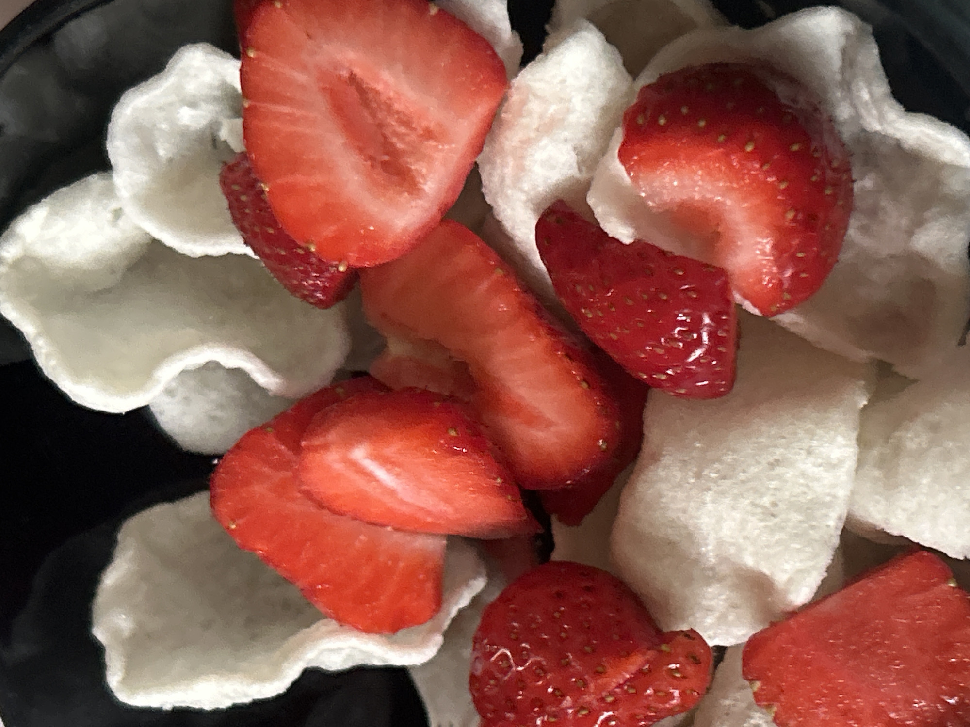 The image depicts a close-up view of a bowl of cut strawberries mixed with white, puffed rice crackers. The strawberries are bright red and appear fresh, with their seeds visible. The crackers are irregular in shape and texture. The bowl itself seems to be dark, possibly black. The location is Unknown, Unknown.