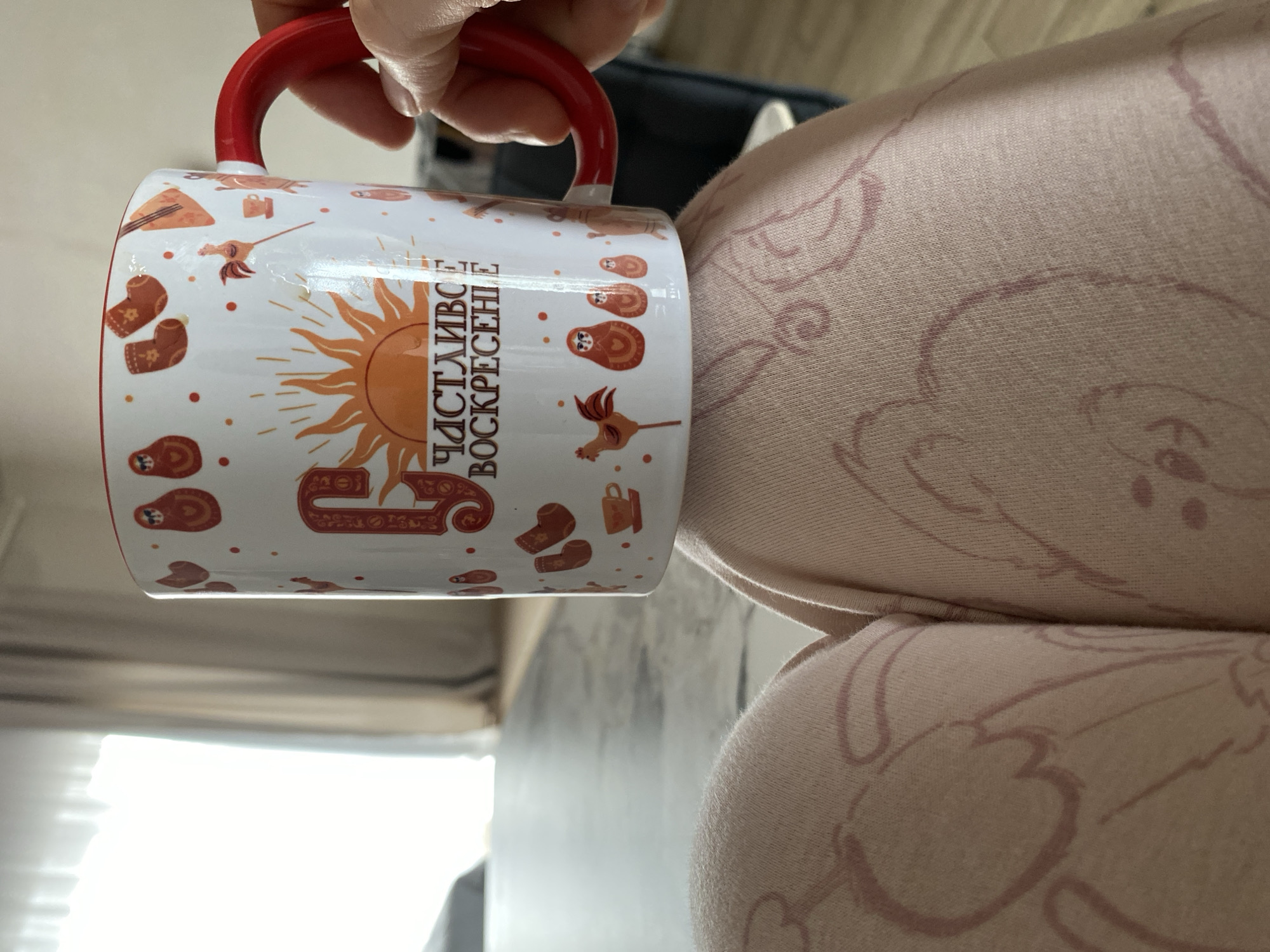 A hand is holding a white mug with a red handle and rim. The mug is decorated with various orange and brown folk art-style images including nesting dolls, slippers, and a sun. Vertical text in Cyrillic is visible on the mug. The mug is being held against a background of soft pink fabric with a repeating pattern of outlined teddy bears. A blurred, pale-colored wall and a window with bright light are visible in the background. The location is potentially in the city of Chaikovsky, Russia, implied but not confirmed by the design on the mug.